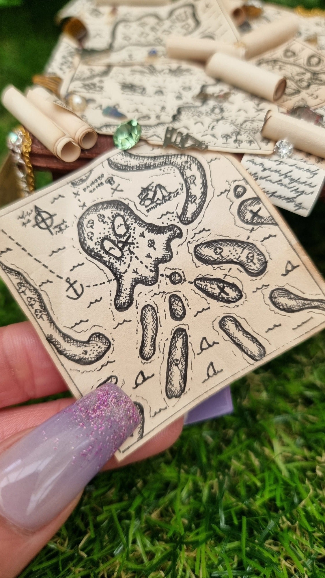 Hand holds a tiny 1:12 scale treasure map that looks like a kraken attached to a 1:12 scale dollshouse brown table scattered with pirate treasure maps, bottle of rum and treasure.