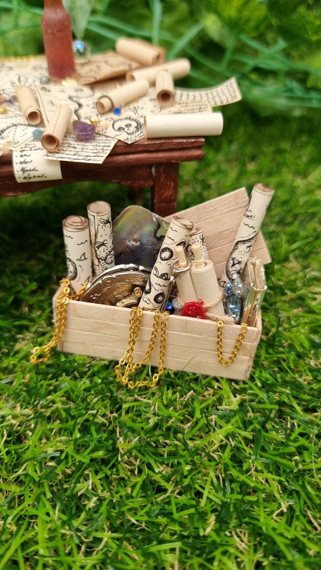 1:12 scale dollshouse brown table scattered with pirate treasure maps, bottle of rum and treasure. Wooden crate filled with treasure sits below the table, filled with plates, maps, chains and scroll