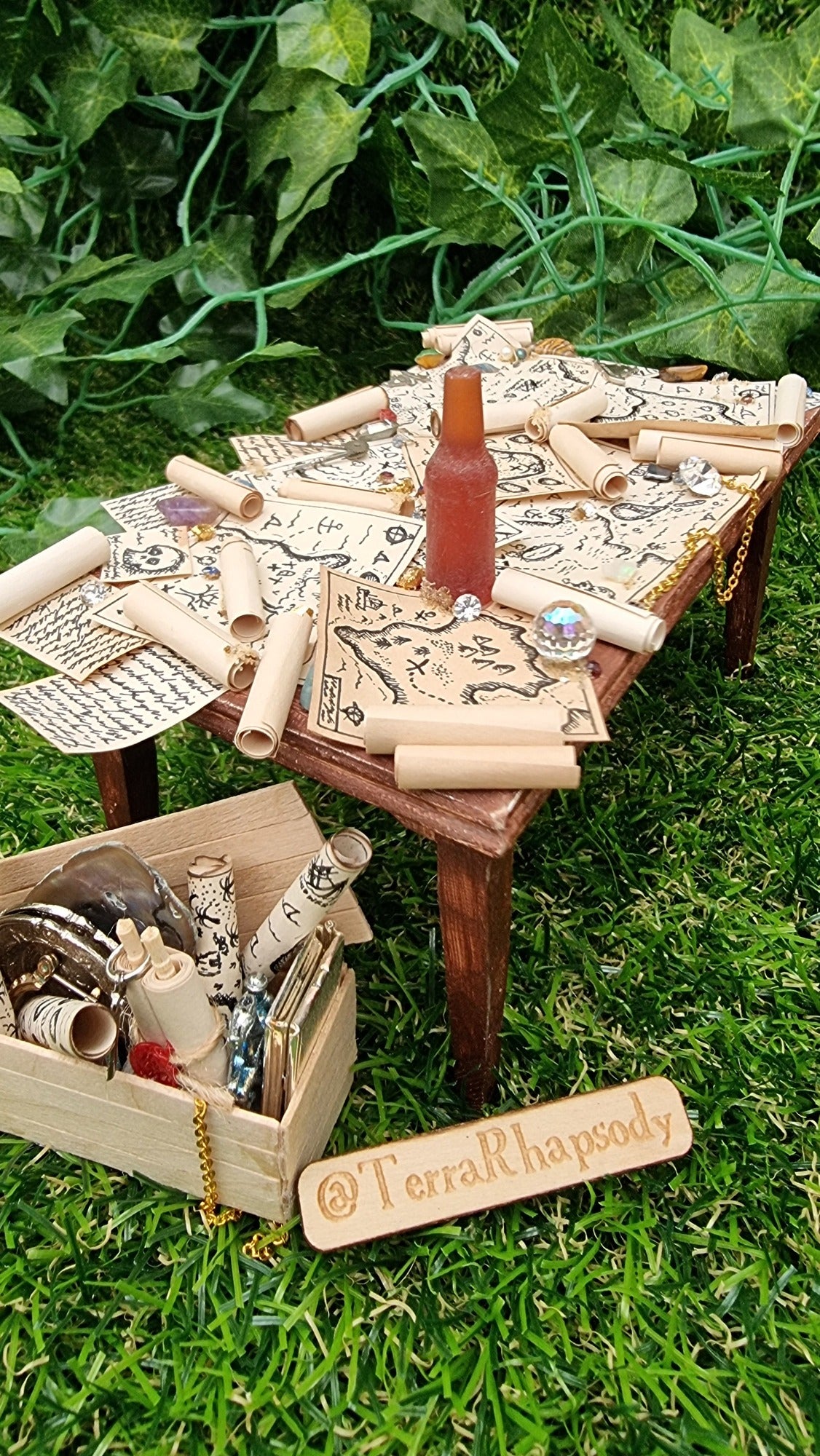 1:12 scale dollshouse brown table scattered with pirate treasure maps, bottle of rum and treasure. Wooden crate filled with treasure sits below the table and @TerraRhapsody is written on wood in the foreground