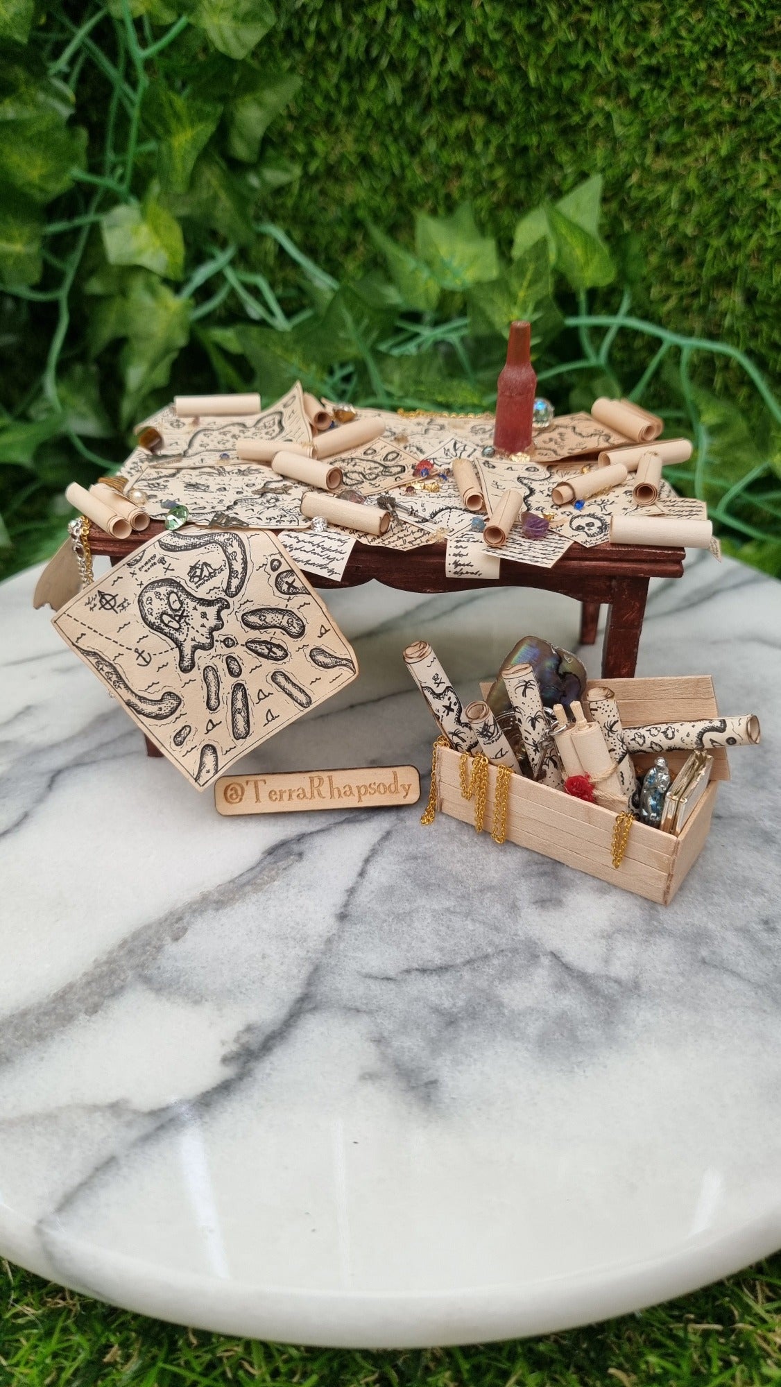 1:12 scale dollshouse brown table scattered with pirate treasure maps, bottle of rum and treasure. Wooden crate filled with treasure sits below the table