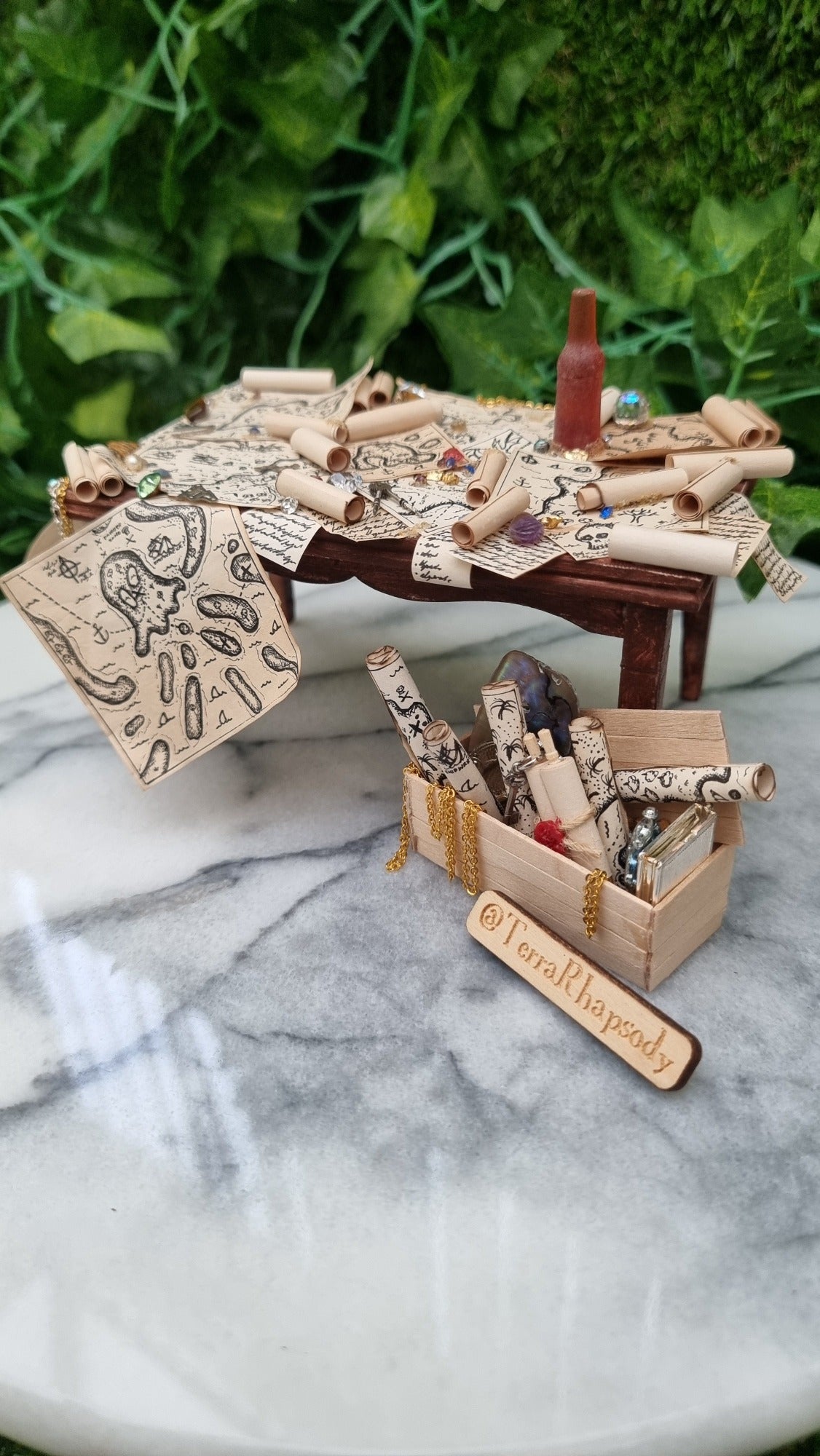 1:12 scale dollshouse brown table scattered with pirate treasure maps, bottle of rum and treasure. Wooden crate filled with treasure sits below the table