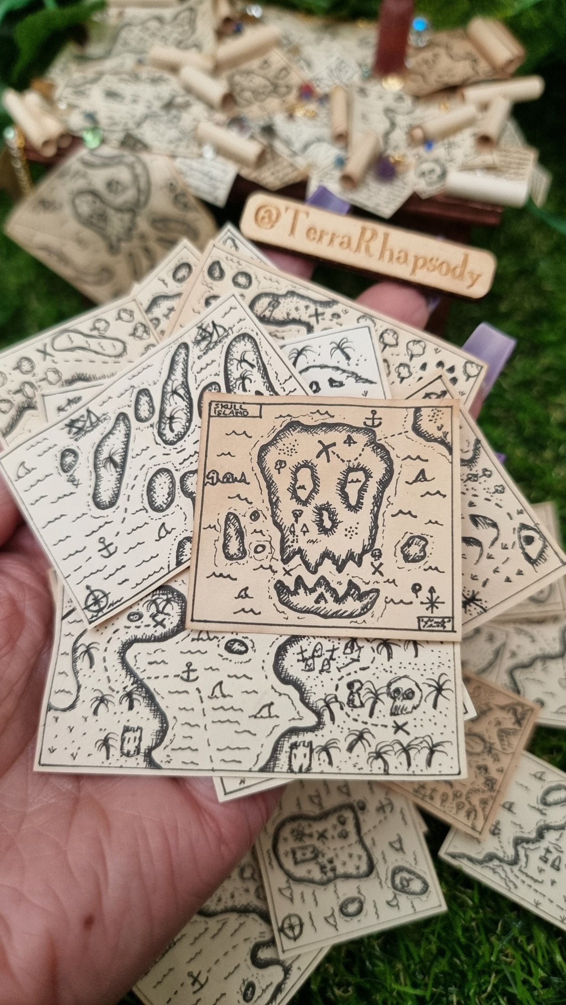 A hand holds a bundle of 1:12 scale beige coloured miniature treasure maps, hand-drawn with tiny island, sea and a map to treasure. The top map has an island that look like a skull