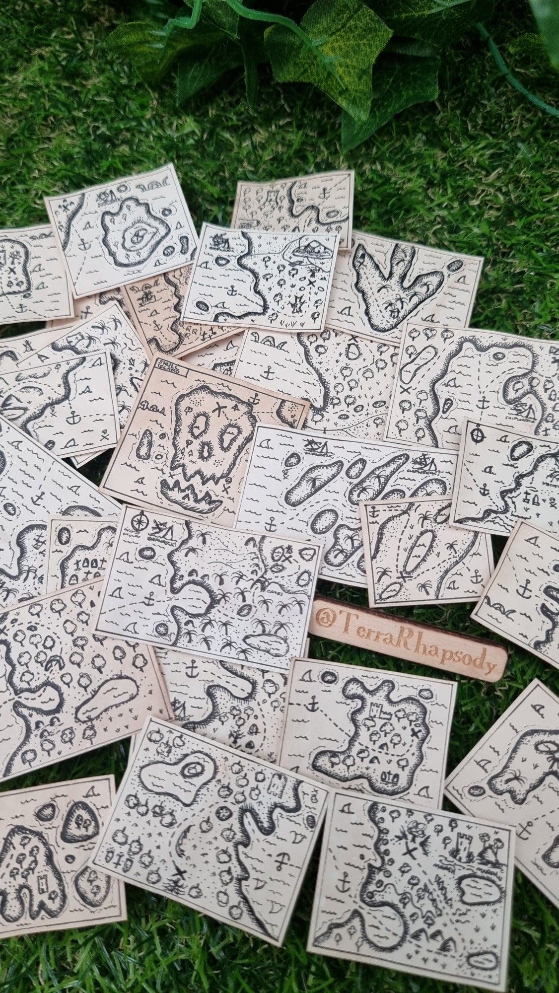 Numerous 1:12 scale miniature treasure maps are arranged around a small sign saying @TerraRhapsody. Each map is different and individually hand drawn with different islands, sea and treasure locations