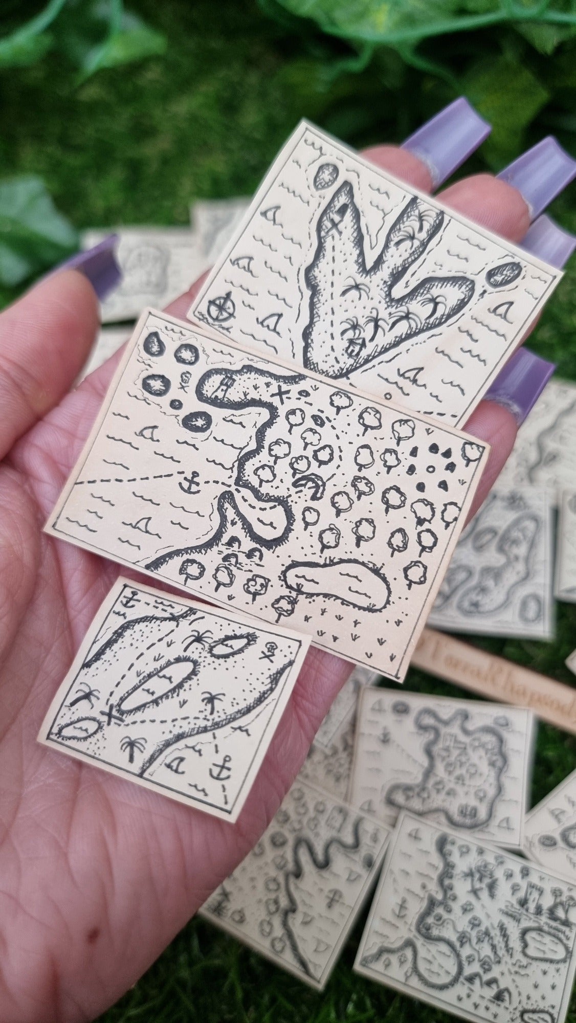  A hand holds a set of 3 1:12 scale beige coloured miniature treasure maps, hand-drawn with tiny island, sea and a map to treasure. The top map has an island that look like a dinosaur foot