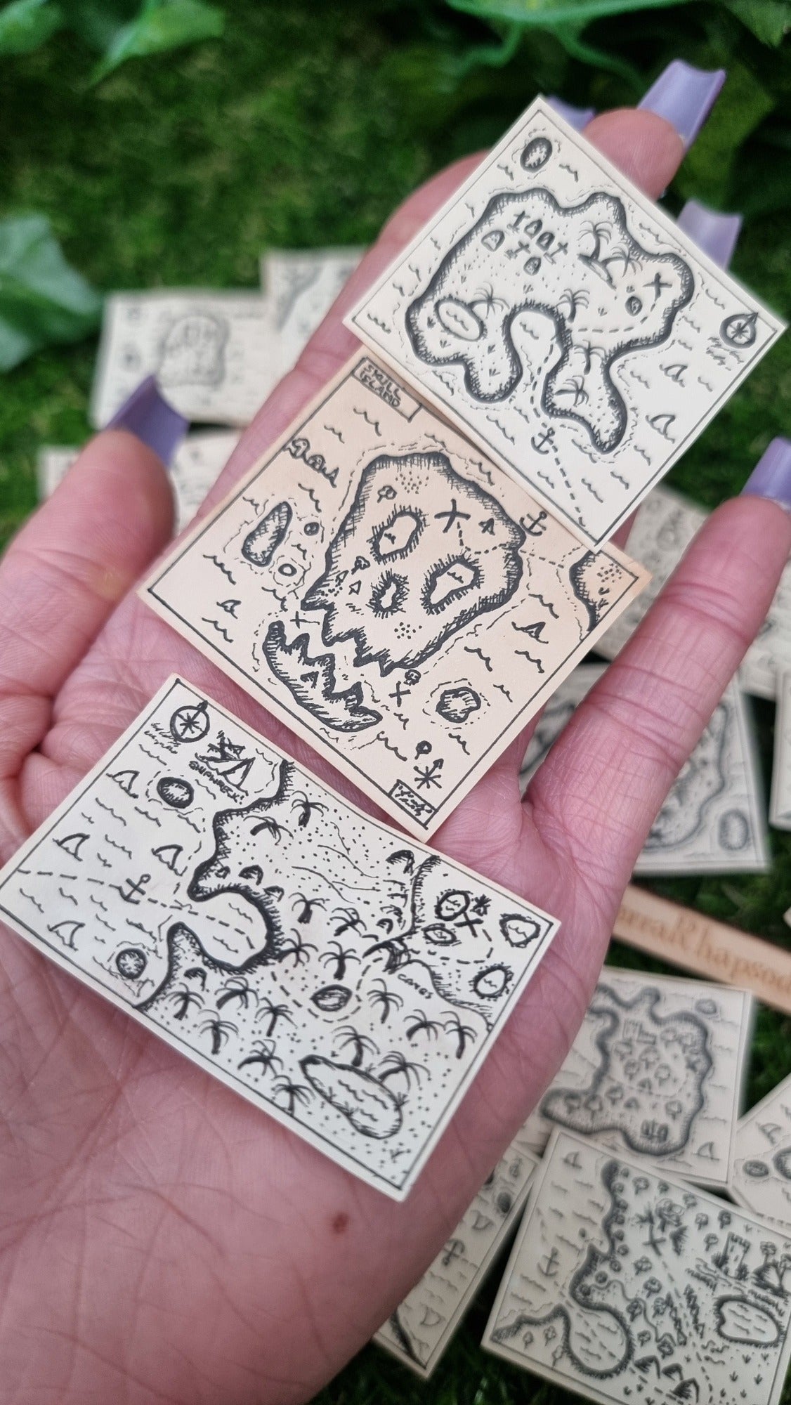 A hand holds a set of 3 1:12 scale beige coloured miniature treasure maps, hand-drawn with tiny island, sea and a map to treasure. One map has an island that look like a skull