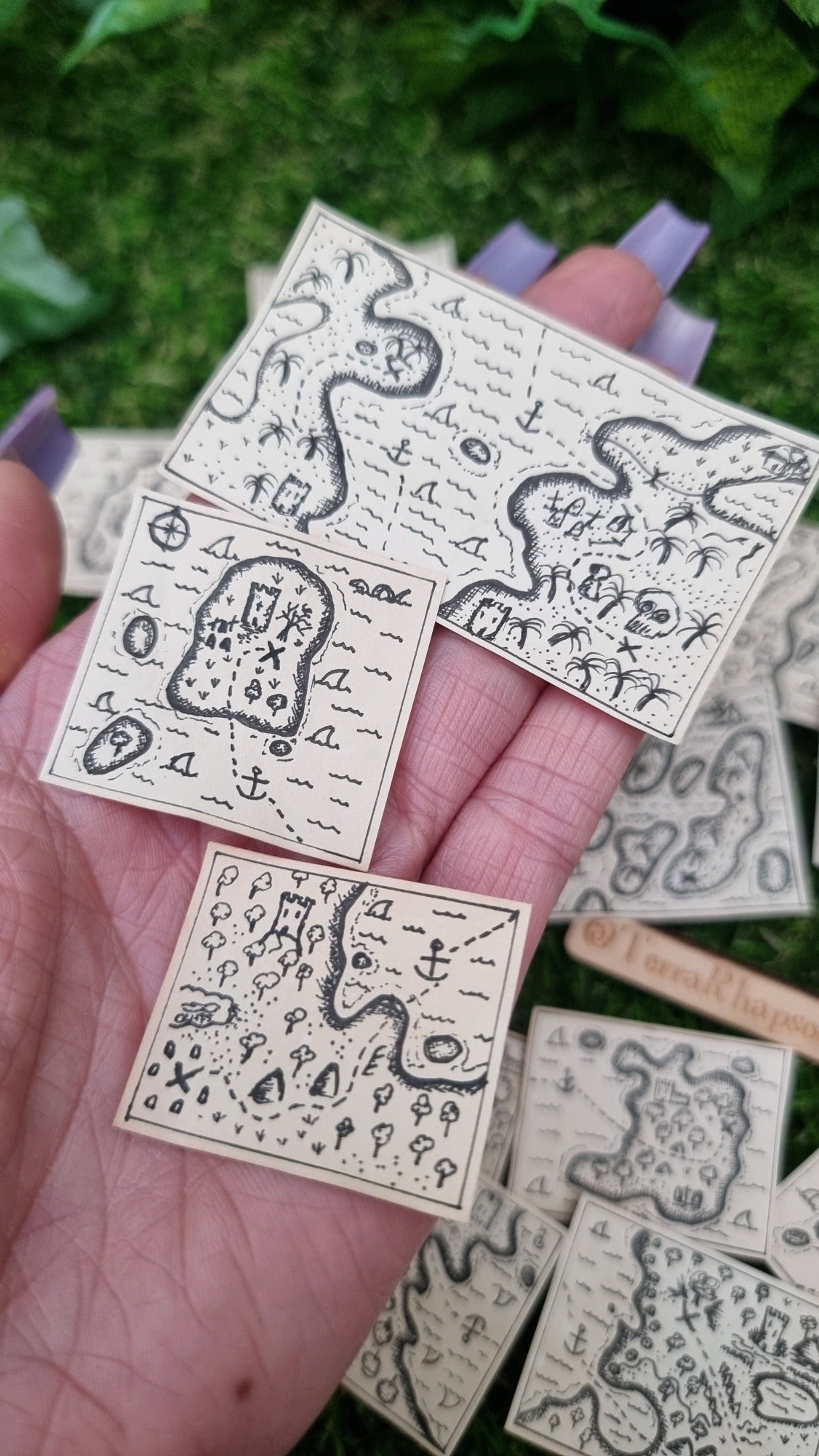 A hand holds a set of 3 1:12 scale beige coloured miniature treasure maps, hand-drawn with tiny island, sea and a map to treasure. The maps have sharks, anchor points and castles