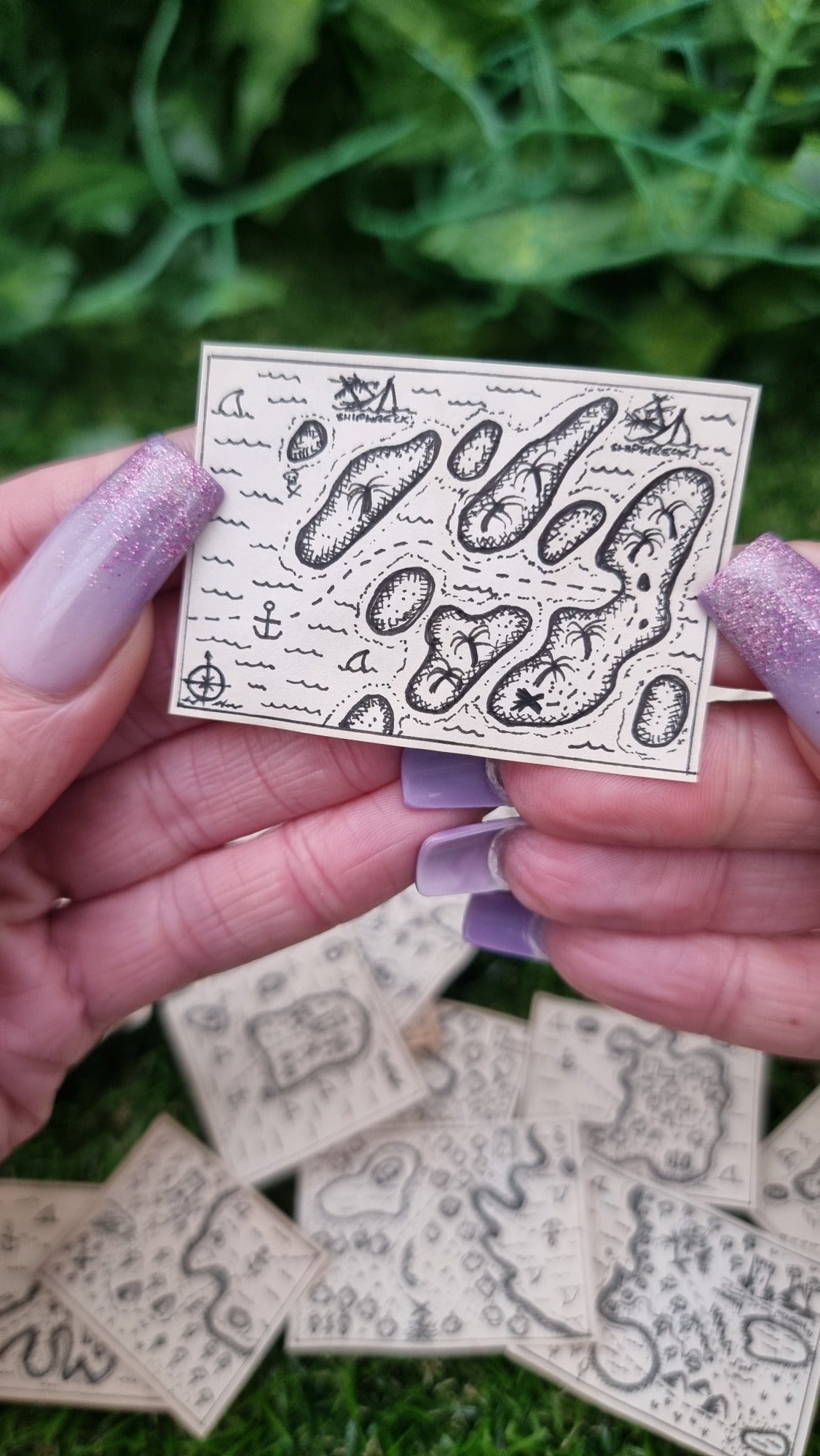 A hand holds a single 1:12 scale beige coloured miniature treasure map. The map has several parallel diagonal islands with a dotted path leading to an 'X' marking the spot. The map has a compass, sharks and 2 shipwrecks