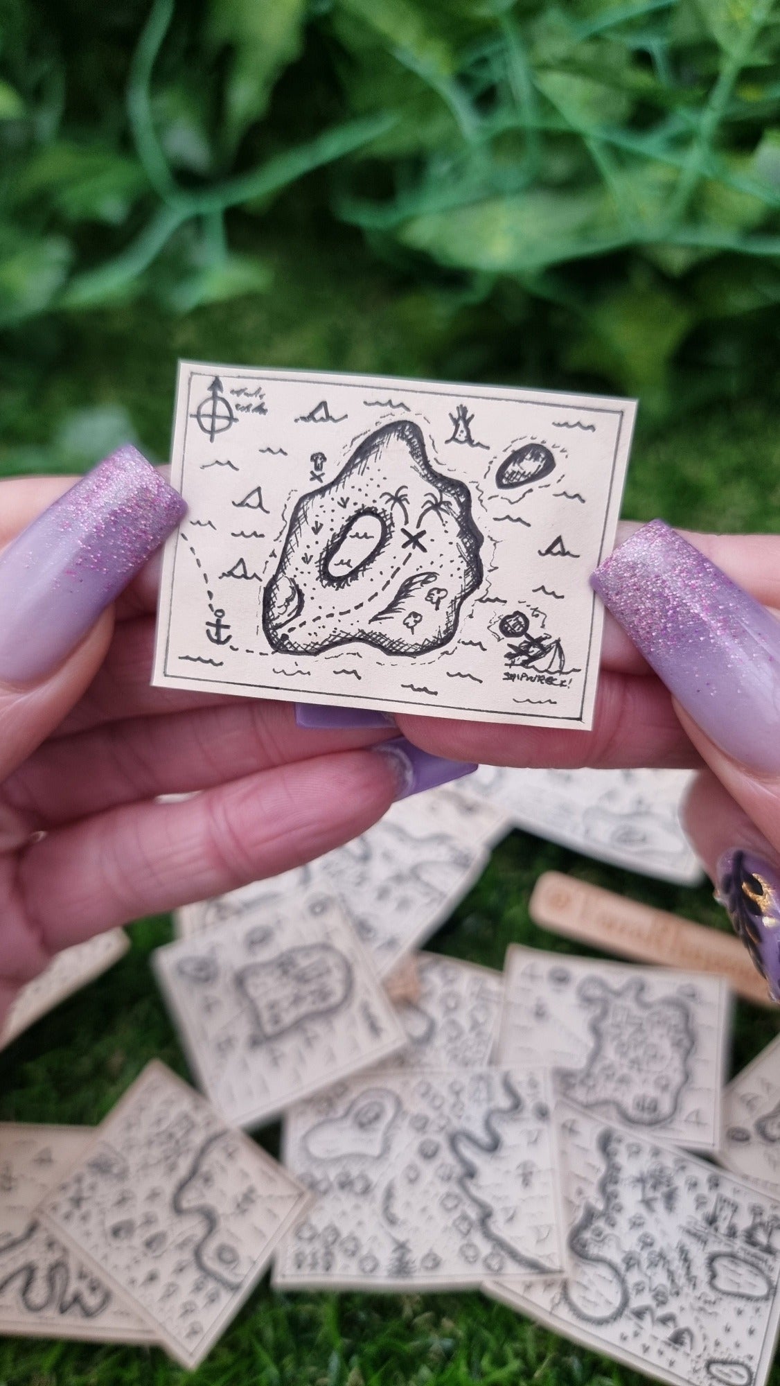  A hand holds a single 1:12 scale beige coloured miniature treasure map, hand-drawn with tiny island, sea and a map to treasure. The island has a small lake, path to the treasure, sharks and shipwreck