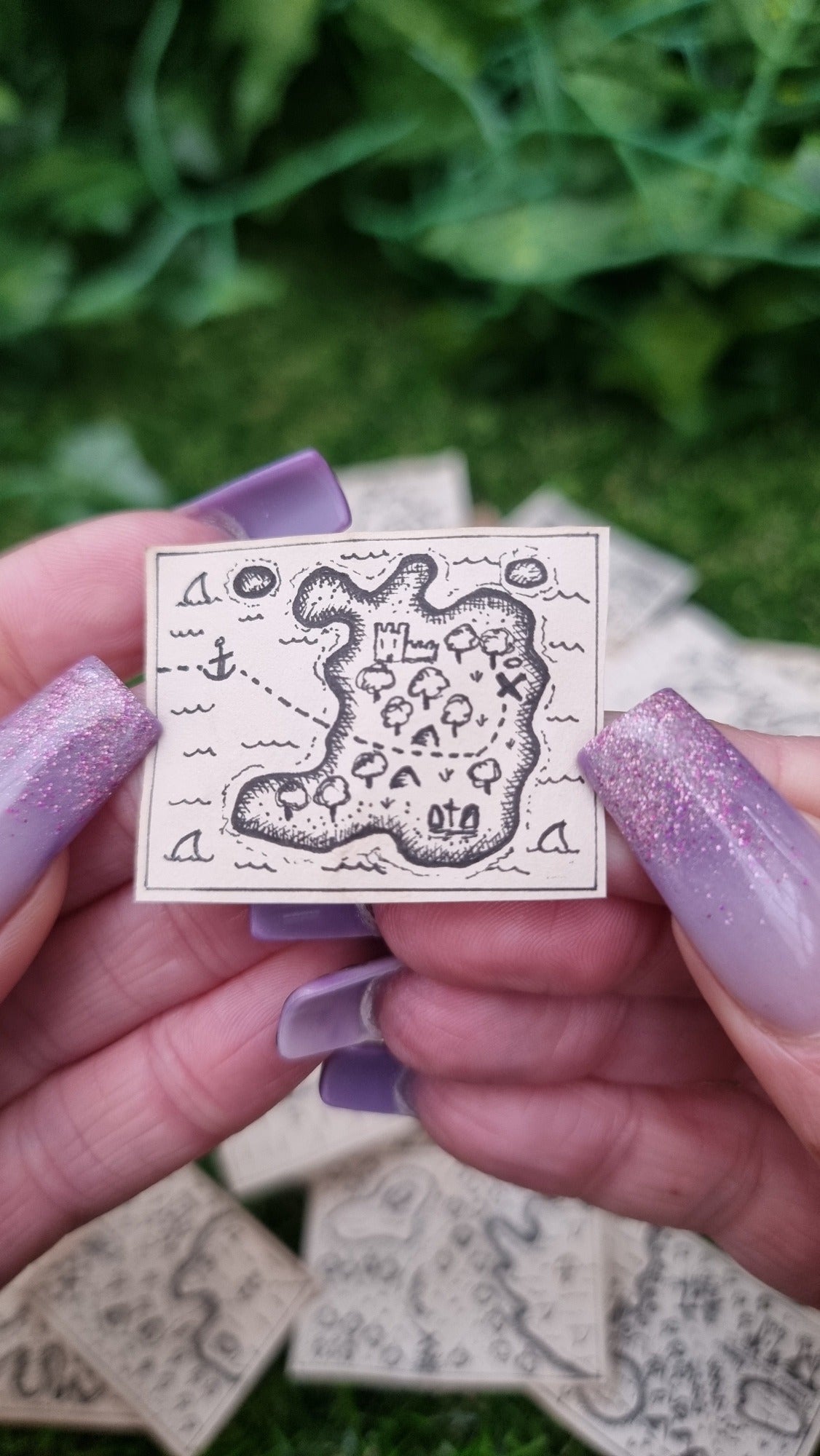 A hand holds a single 1:12 scale beige coloured miniature treasure map, hand-drawn with tiny island, sea and a map to treasure. The island has a forest, graveyard, castle and path to the treasure