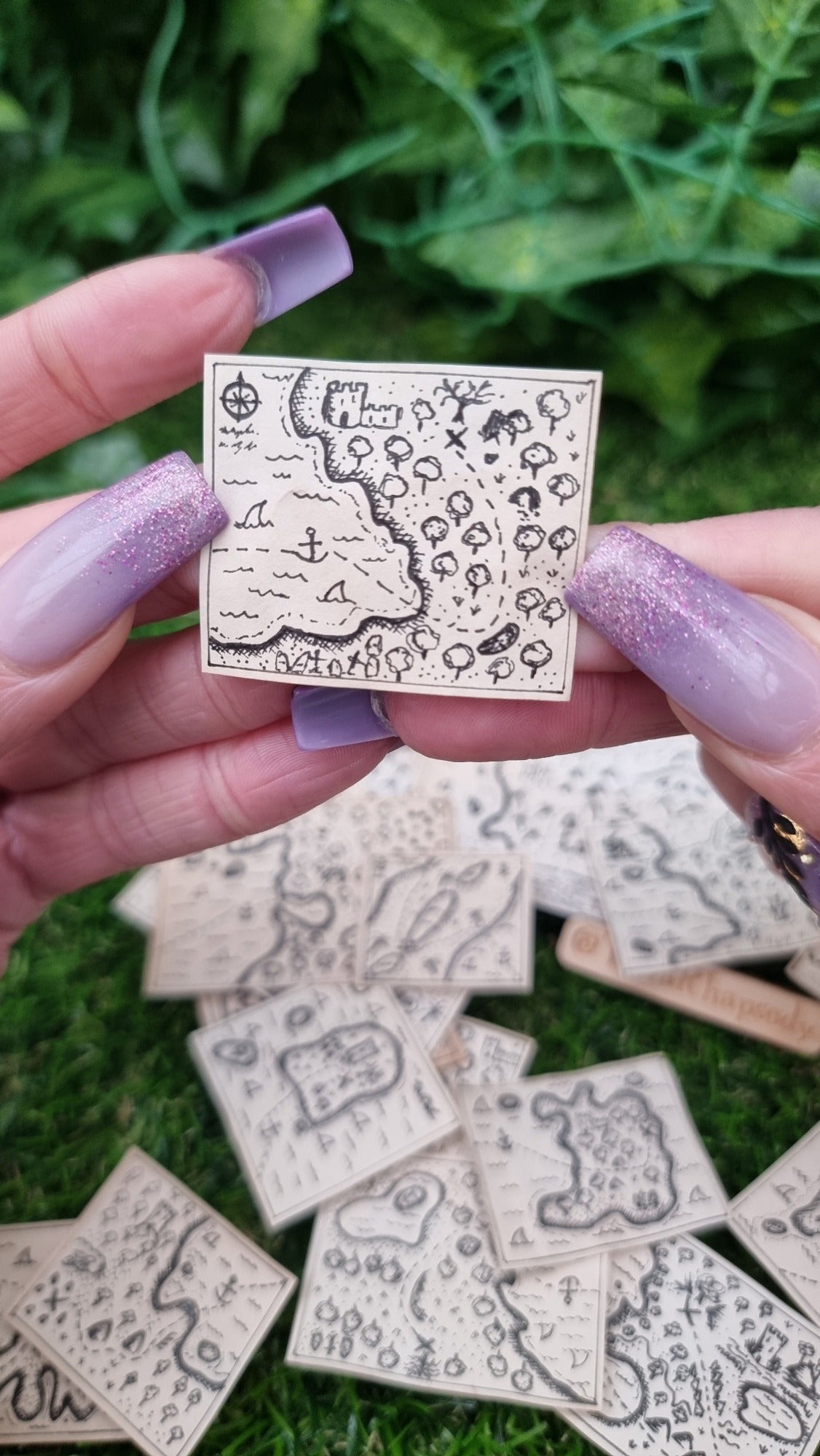 A hand holds a single 1:12 scale beige coloured miniature treasure map, hand-drawn with tiny island, sea and a map to treasure. The island has a forest, graveyard, castle and path to the treasure