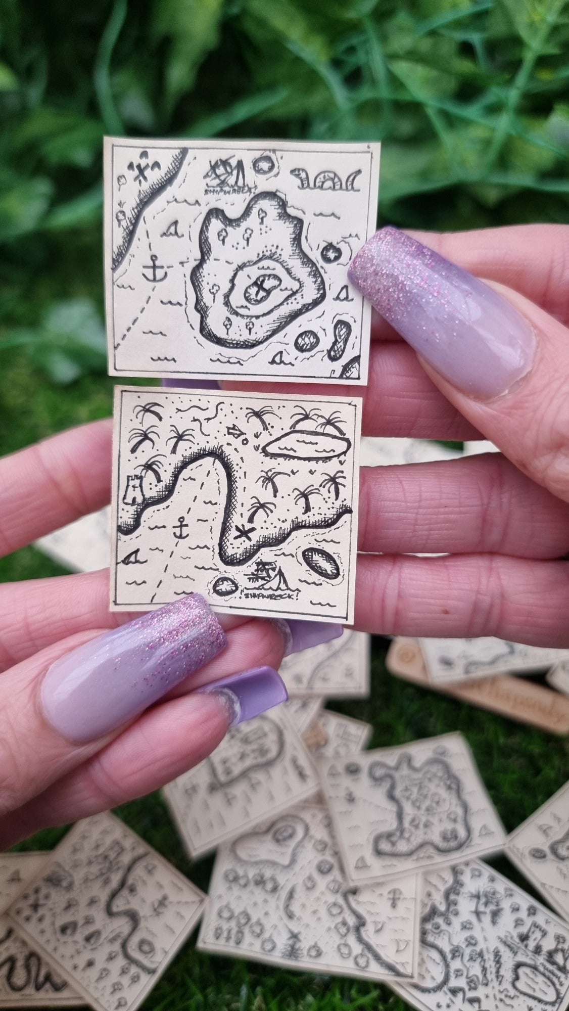 A hand holds 2 1:12 scale beige coloured miniature treasure maps, hand-drawn with tiny islands, sea and a map to treasure. The maps show routes to an 'X' marking the spot, sharks, shipwreck and sea monster