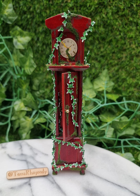 Miniature 1:12 scale grandfather clock sits of a marble slab, covered with climbing green ivy and moss. The clock face is stopped at 1.50 and the door in the body of the clock is ajar. @TerraRhapsody is written on wood beside it