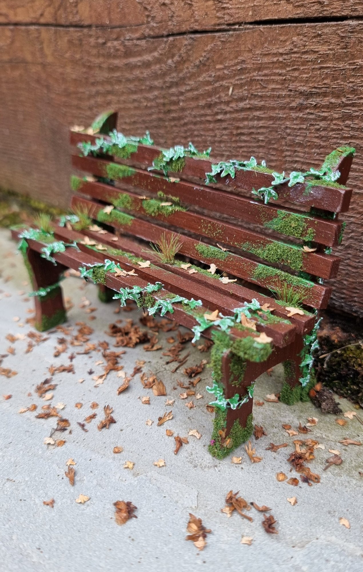 1:12 scale dollhouse wooden bench sits abandoned against a wood and stone background. The bench has green ivy twisting through the slats, dead leaves, grass and moss