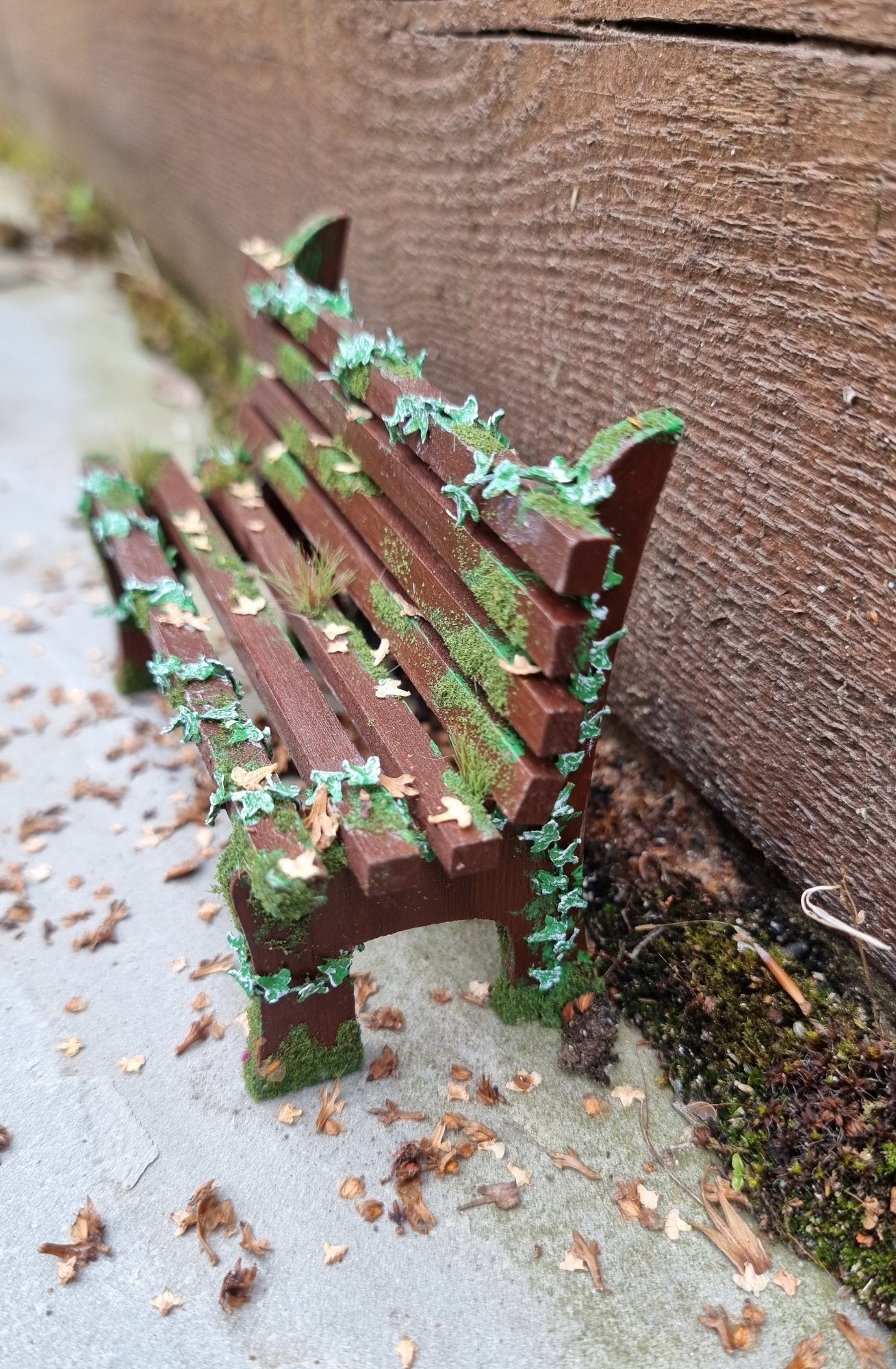 1:12 scale dollhouse wooden bench sits abandoned against a wood and stone background. The bench has green ivy twisting through the slats, dead leaves, grass and moss