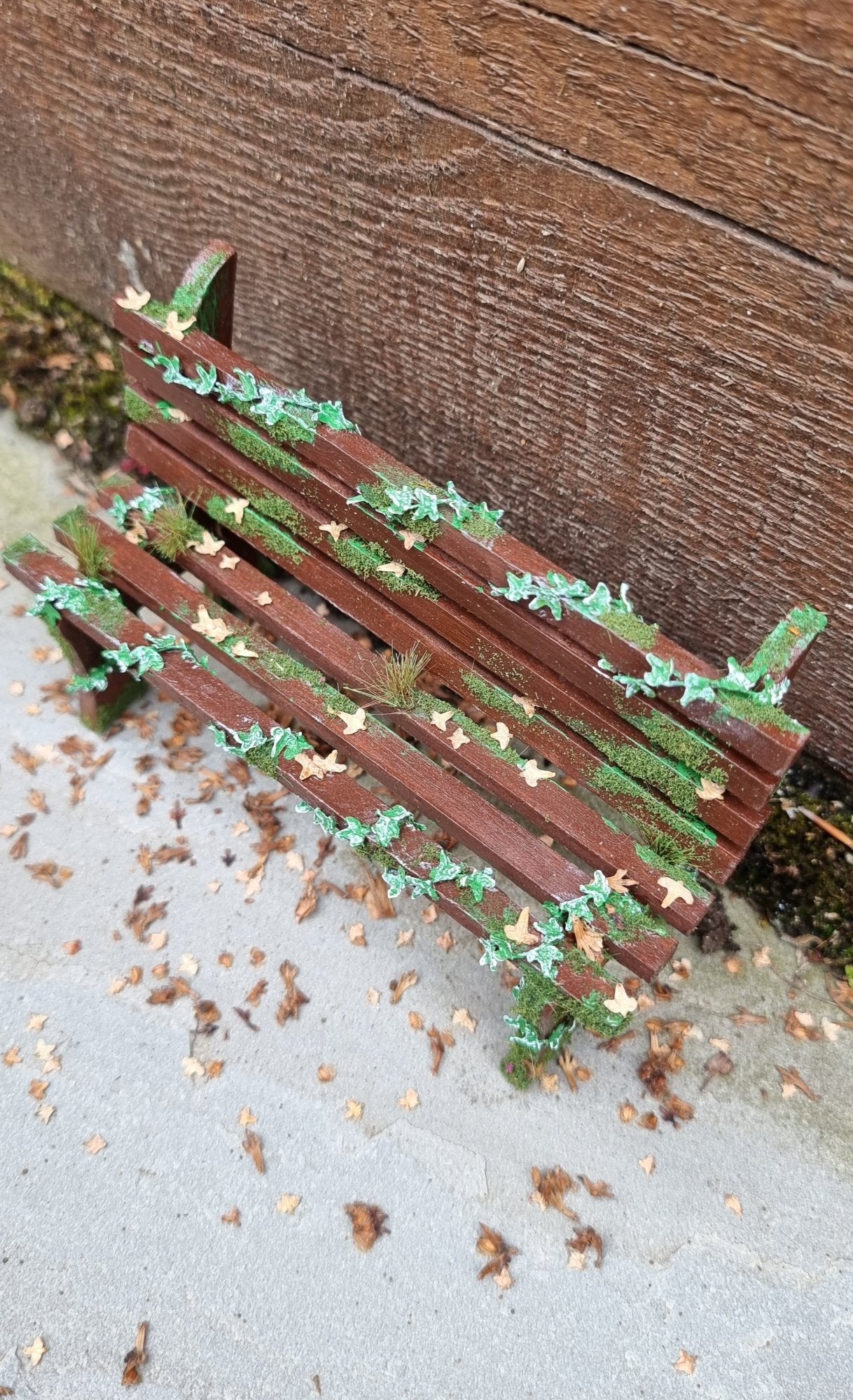 1:12 scale dollhouse wooden bench sits abandoned against a wood and stone background. The bench has green ivy twisting through the slats, dead leaves, grass and moss