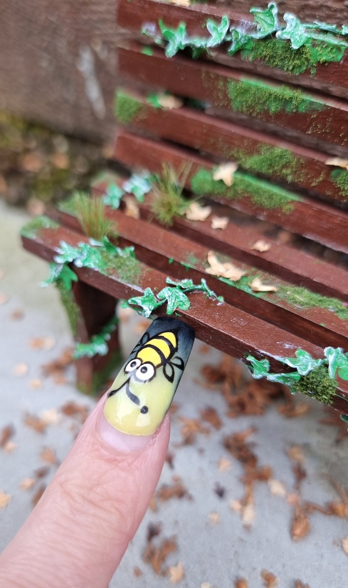 1:12 scale dollhouse wooden bench sits abandoned against a wood and stone background. The bench has green ivy twisting through the slats, dead leaves, grass and moss. A finger points for scale