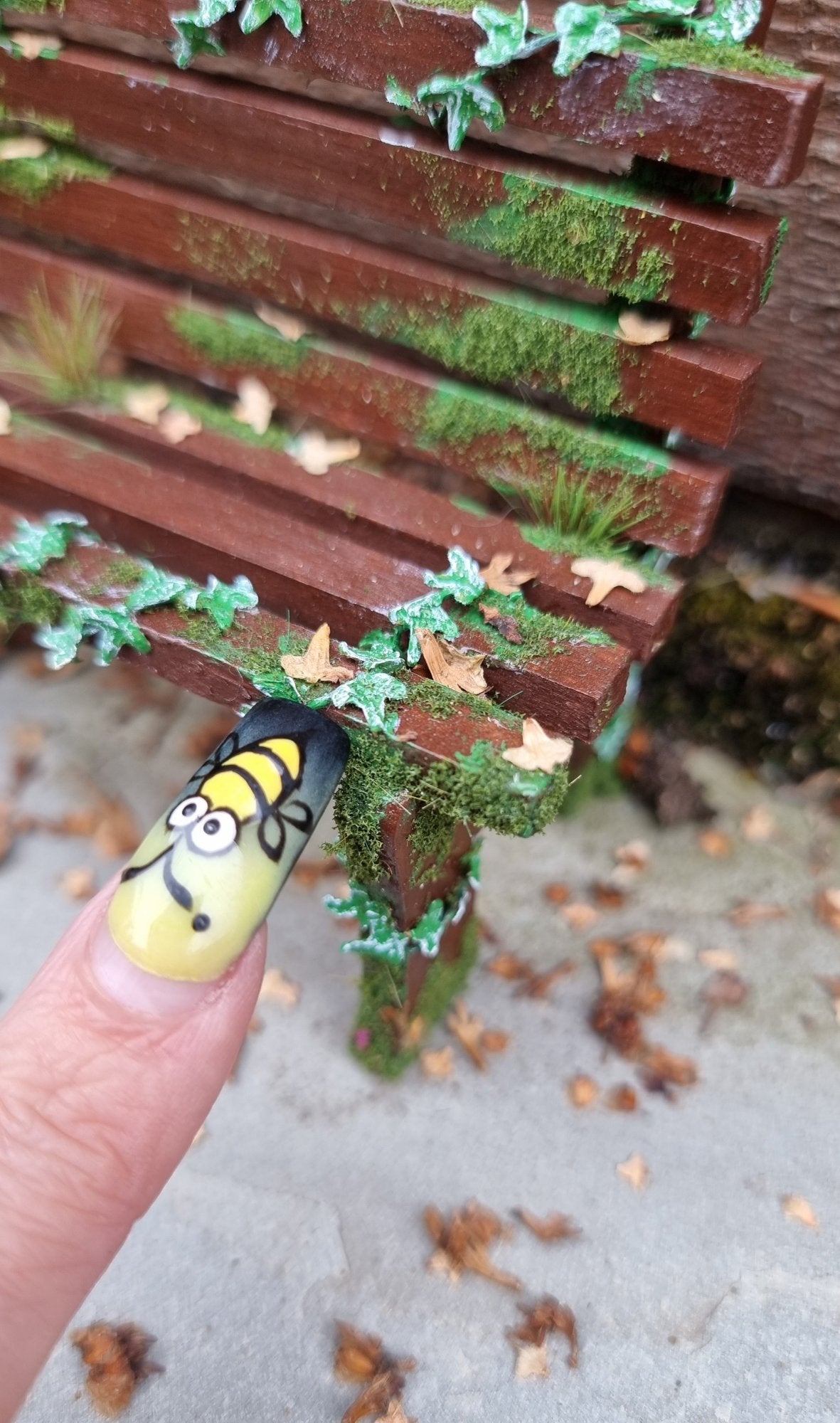 1:12 scale dollhouse wooden bench sits abandoned against a wood and stone background. The bench has green ivy twisting through the slats, dead leaves, grass and moss. A finger points for scale