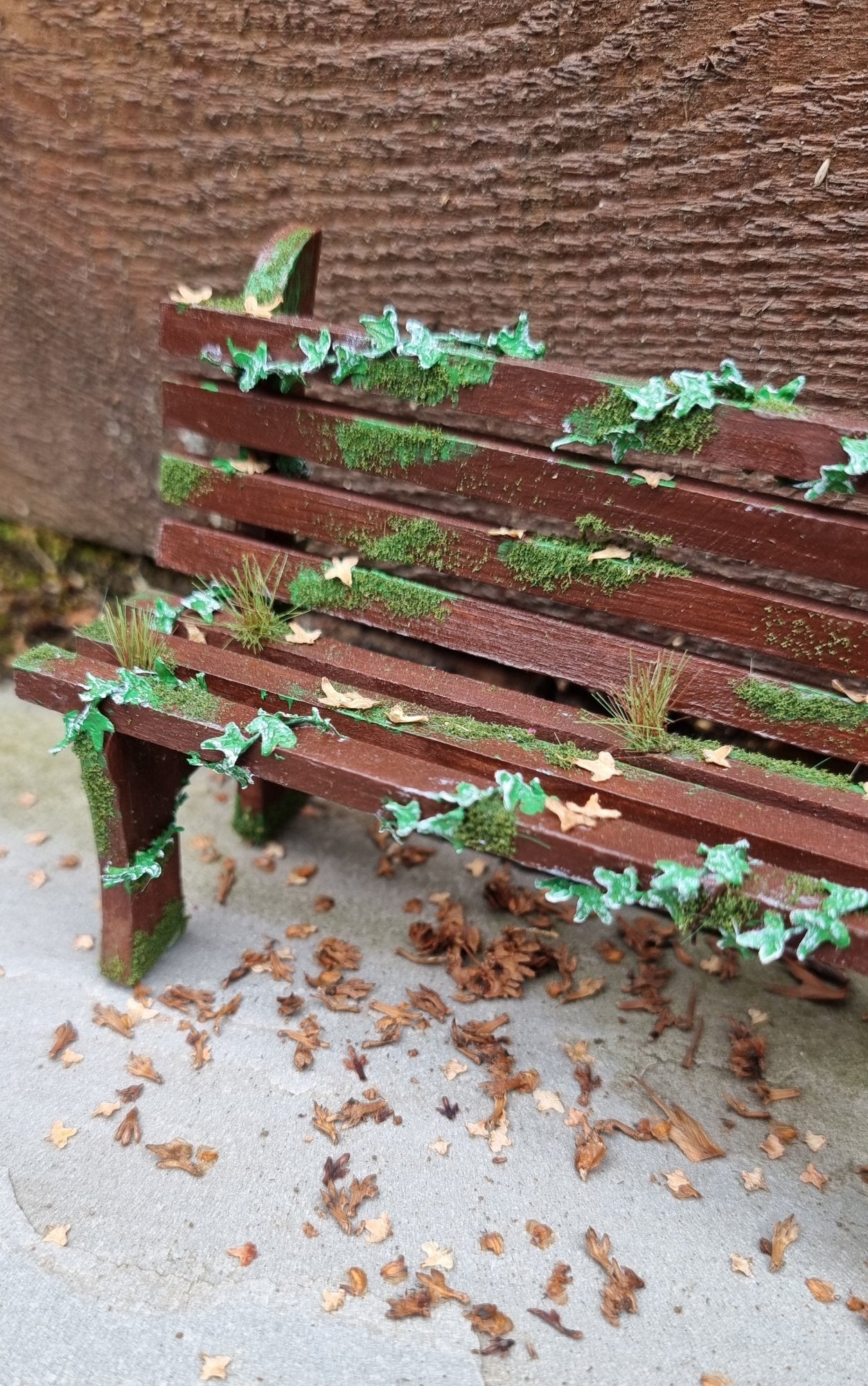 1:12 scale dollhouse wooden bench sits abandoned against a wood and stone background. The bench has green ivy twisting through the slats, dead leaves, grass and moss