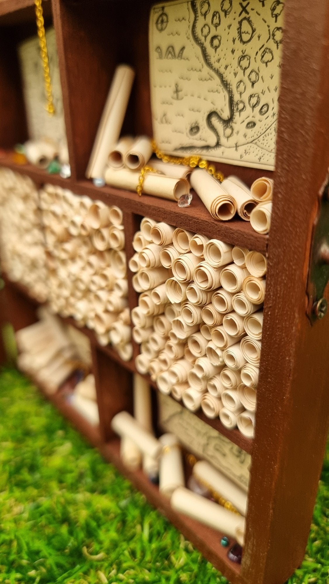 Detail of a miniature 1:12 scale tall brown cabinet filled with scrolls of paper sitting against a green background. The cabinet shows alcoves filled with parchment rolls, with surrounding alcoves filled with vintage pirate maps, parchment rolls, chains and gemstones.