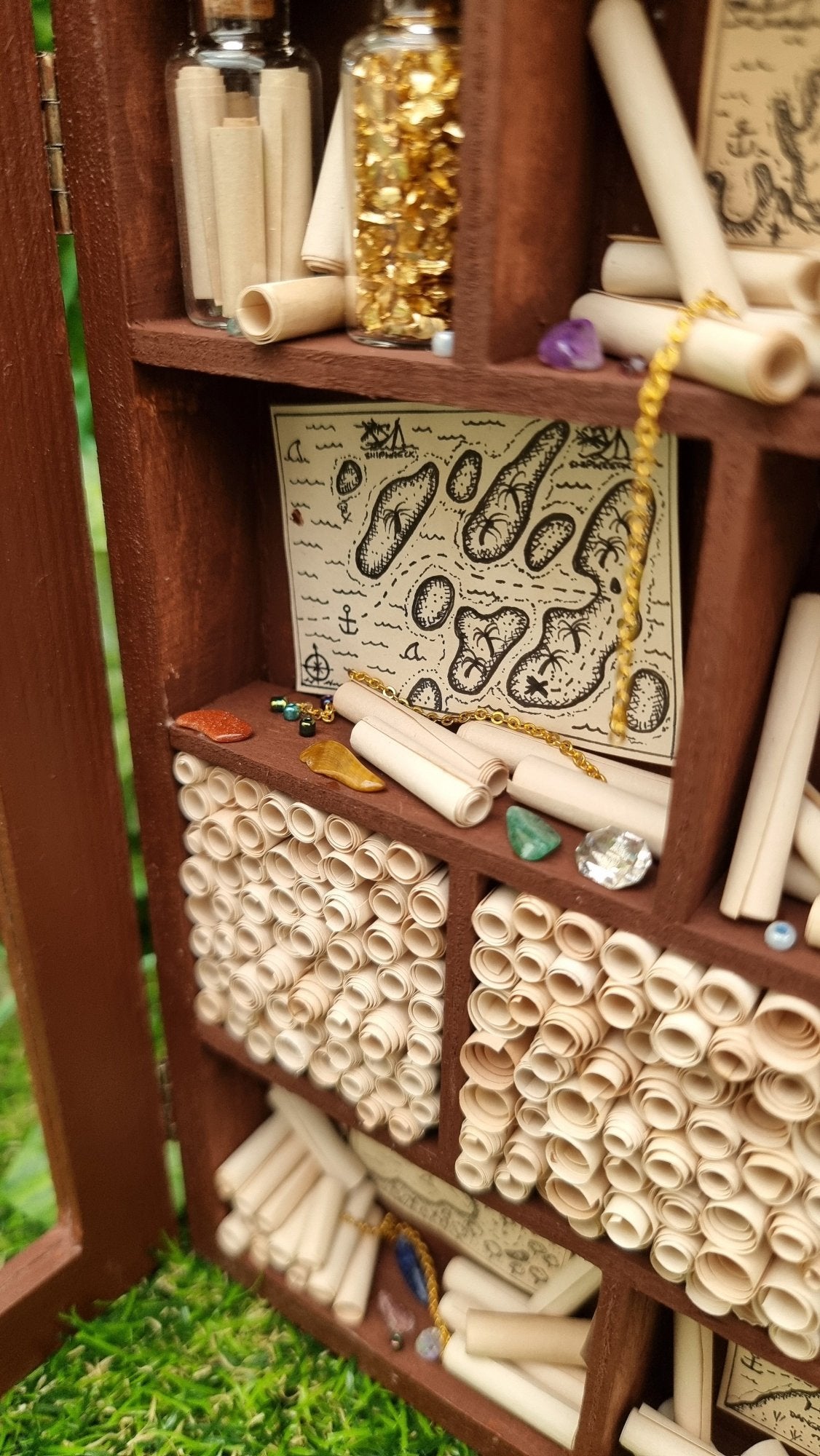 Detail of a miniature 1:12 scale tall brown cabinet filled with scrolls of paper sitting against a green background. The cabinet shows one alcove with a vintage pirate map pinned up, surrounded by rolls of parchment, chain and gemstones. Surrounding alcoves are fiilled with rolls of parchment and glass bottles