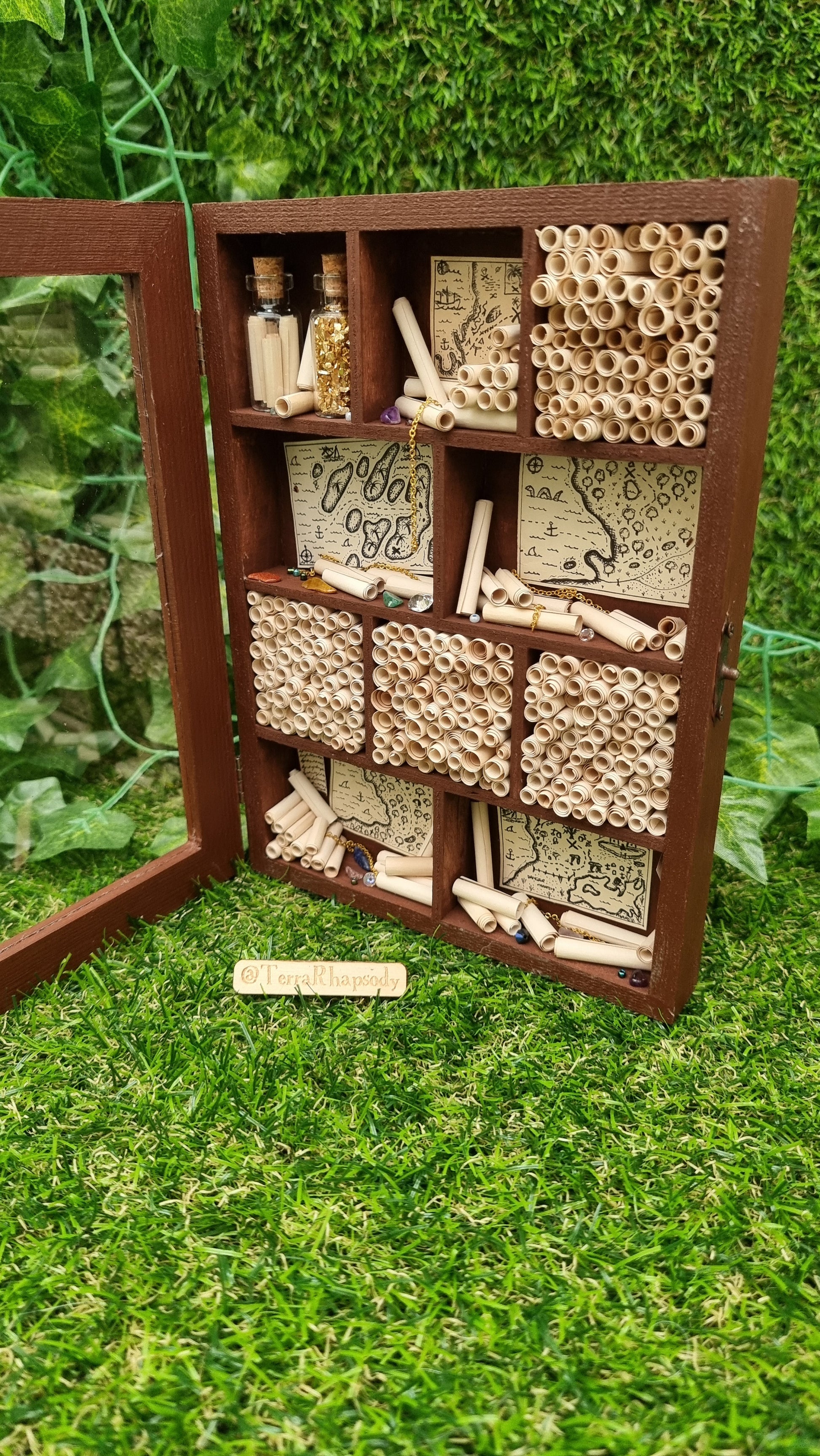 A miniature 1:12 scale tall brown cabinet filled with scrolls of paper sits against a green background. The cabinet has a clear front and 10 shelves or alcoves filled with treasures. The shelves contain vintage pirate maps, glass vials, chains and gemstones. @Terra Rhapsody is written on wood in front of it