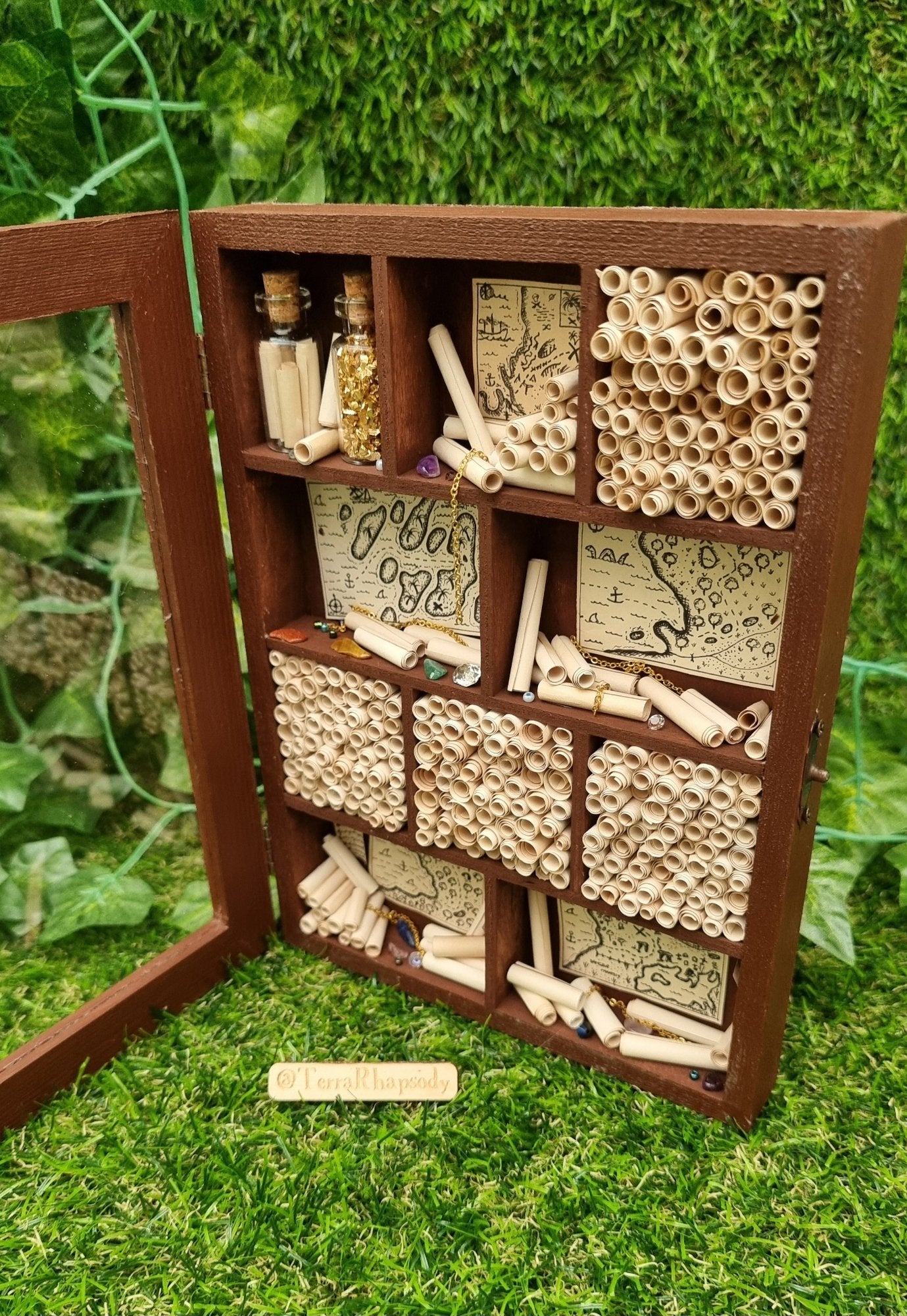 A miniature 1:12 scale tall brown cabinet filled with scrolls of paper sits against a green background. The cabinet has a clear front and 10 shelves or alcoves filled with treasures. The shelves contain vintage pirate maps, glass vials, chains and gemstones. @Terra Rhapsody is written on wood in front of it