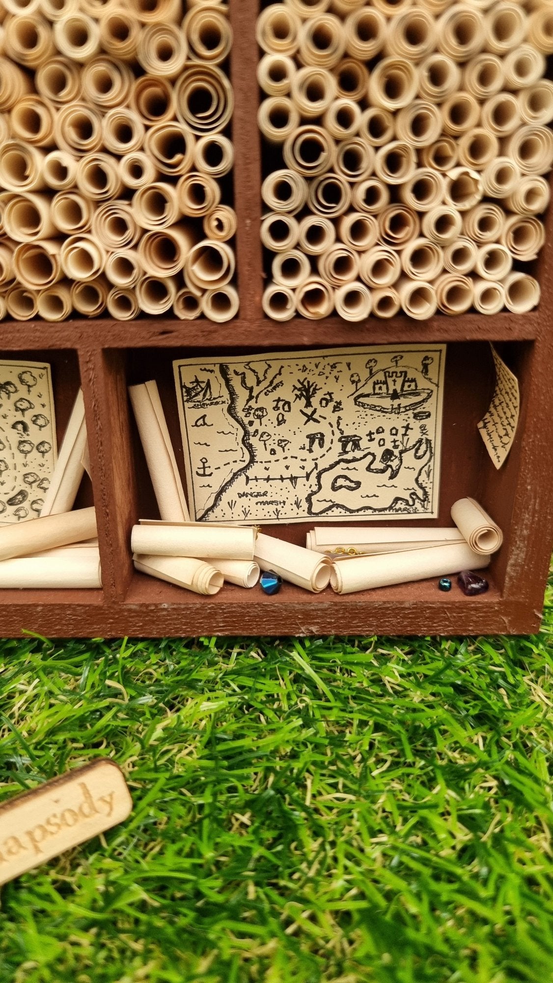 Detail of a miniature 1:12 scale tall brown cabinet filled with scrolls of paper sitting against a green background. The cabinet shows one alcove with a vintage pirate map pinned up and rolls of parchment and gemstones. Surrounding alcoves are fiilled with rolls of parchment