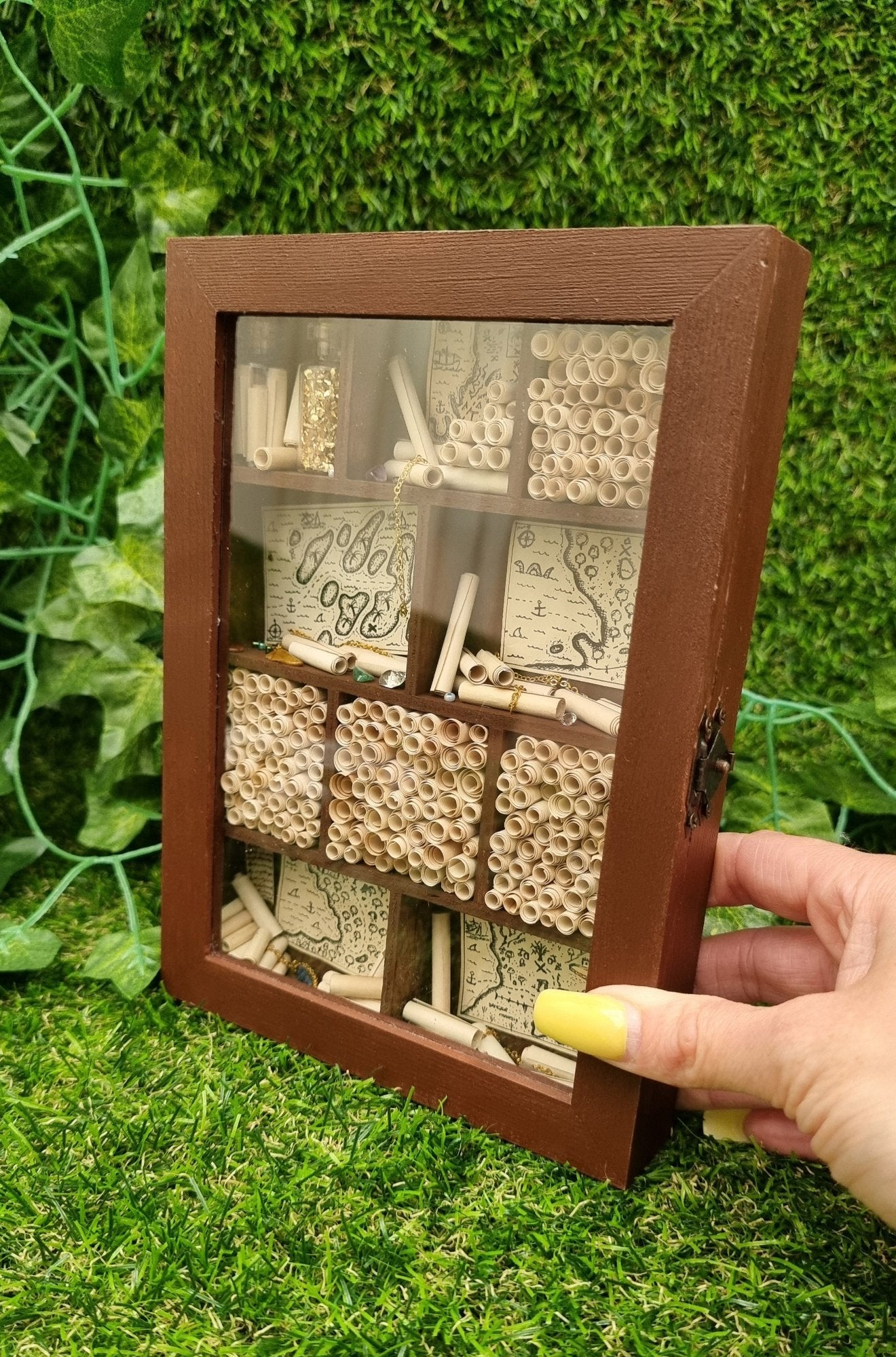 A miniature 1:12 scale tall brown cabinet filled with scrolls of paper sits against a green background. The cabinet is closed but has a clear front and 10 shelves or alcoves filled with treasures. The shelves contain vintage pirate maps, glass vials, chains and gemstones. 