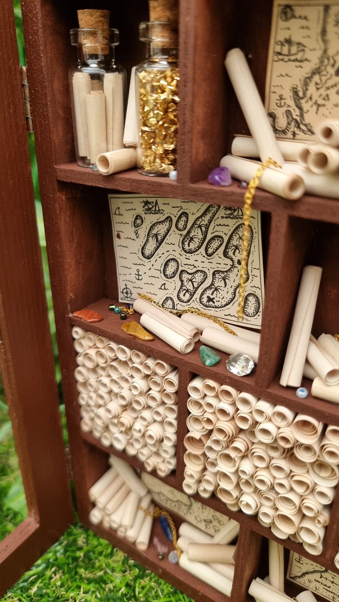 Detail shown of a miniature 1:12 scale tall brown cabinet filled with scrolls of paper sits against a green background. The cabinet has a clear front and 10 shelves or alcoves filled with treasures. The shelves contain vintage pirate maps, glass vials, chains and gemstones