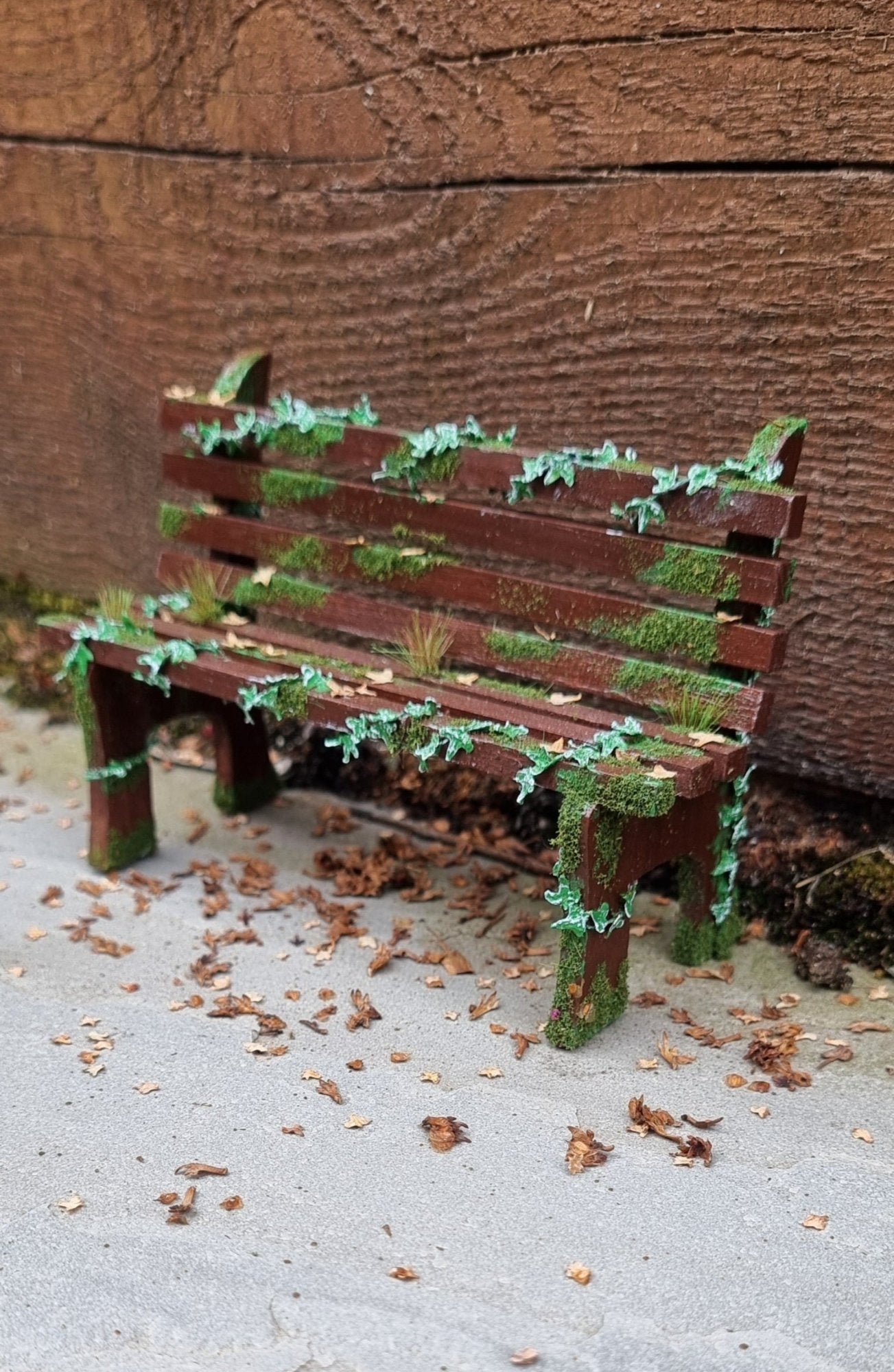 1:12 scale dollhouse wooden bench sits abandoned against a wood and stone background. The bench has green ivy twisting through the slats, dead leaves, grass and moss