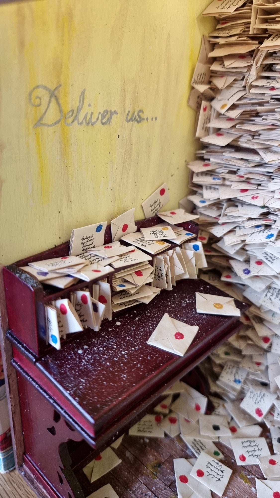 A 1:12 scale miniature desk covered in letters sits against a yellow wall with silver cursive text reading "Deliver Us" above. To the right are shelves crammed with letters. 