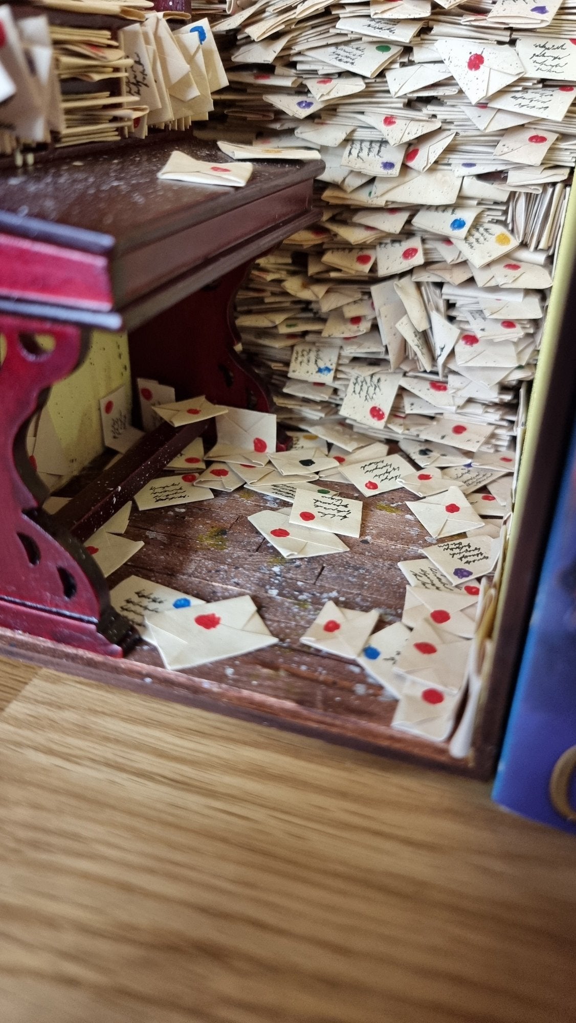 A 1:12 scale miniature book nook diorama is shown from the wooden boarded floor, covered in letters. The back of the diorama are shelves crammed with letters.