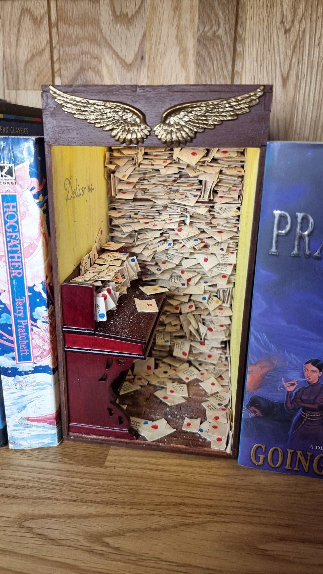 A miniature book nook diorama sits on a shelf beside a copy of Terry Pratchett's Going Postal. The book nook contains a desk littered with letters. Shelves crammed with letters reach from the floor to the roof. The top of the book nook is framed with 2 golden stamped wings