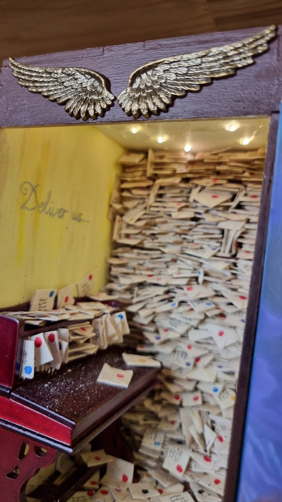 A miniature book nook diorama sits on a shelf beside a copy of Terry Pratchett's Going Postal. The book nook contains a desk littered with letters. Shelves crammed with letters reach from the floor to the roof. The top of the book nook is framed with 2 golden stamped wings. Above the desk on the yellow wall is silver cursive writing saying " Deliver us"