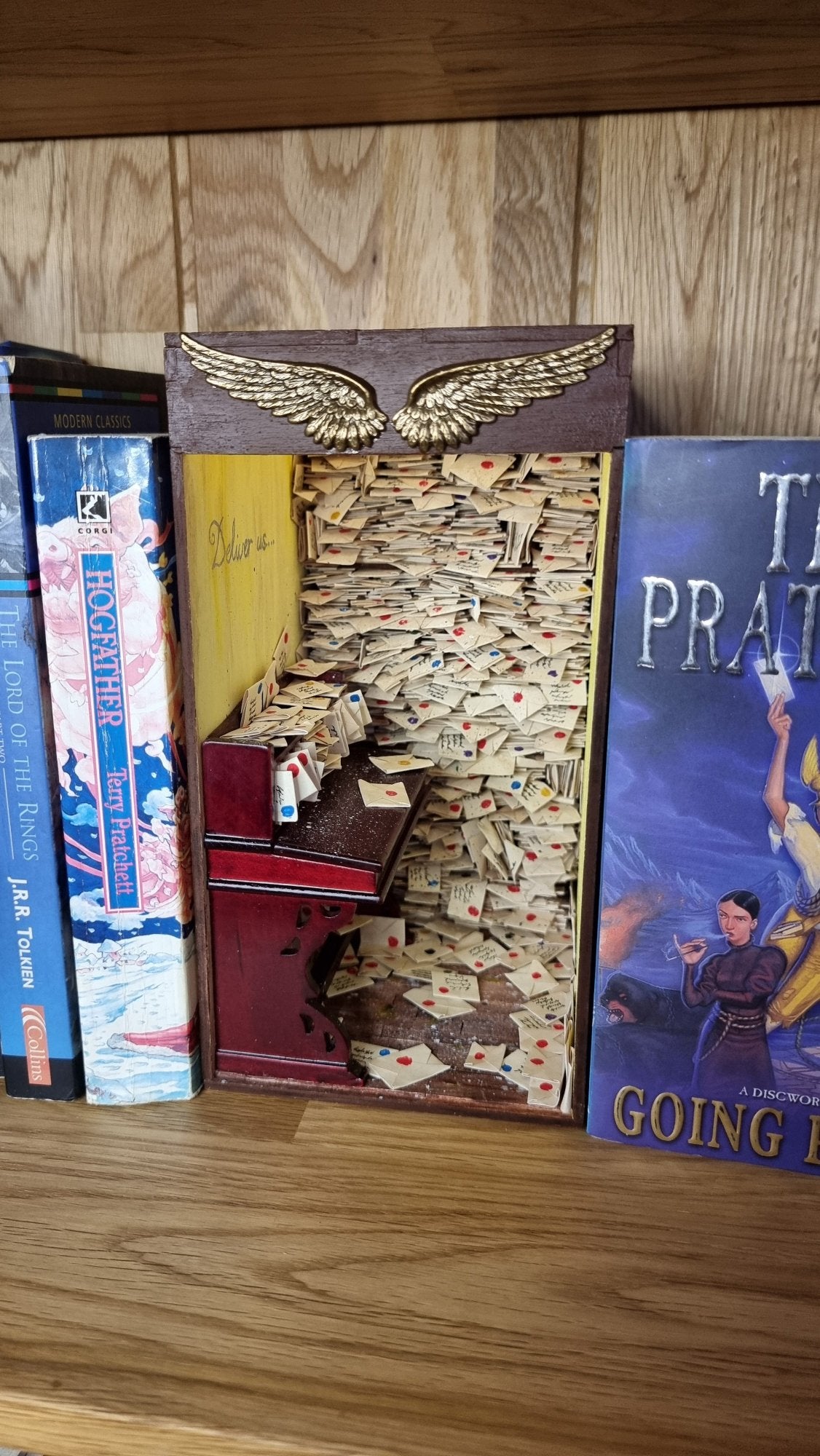 A miniature book nook diorama sits on a shelf beside a copy of Terry Pratchett's Going Postal. The book nook contains a desk littered with letters. Shelves crammed with letters reach from the floor to the roof. The top of the book nook is framed with 2 golden stamped wings