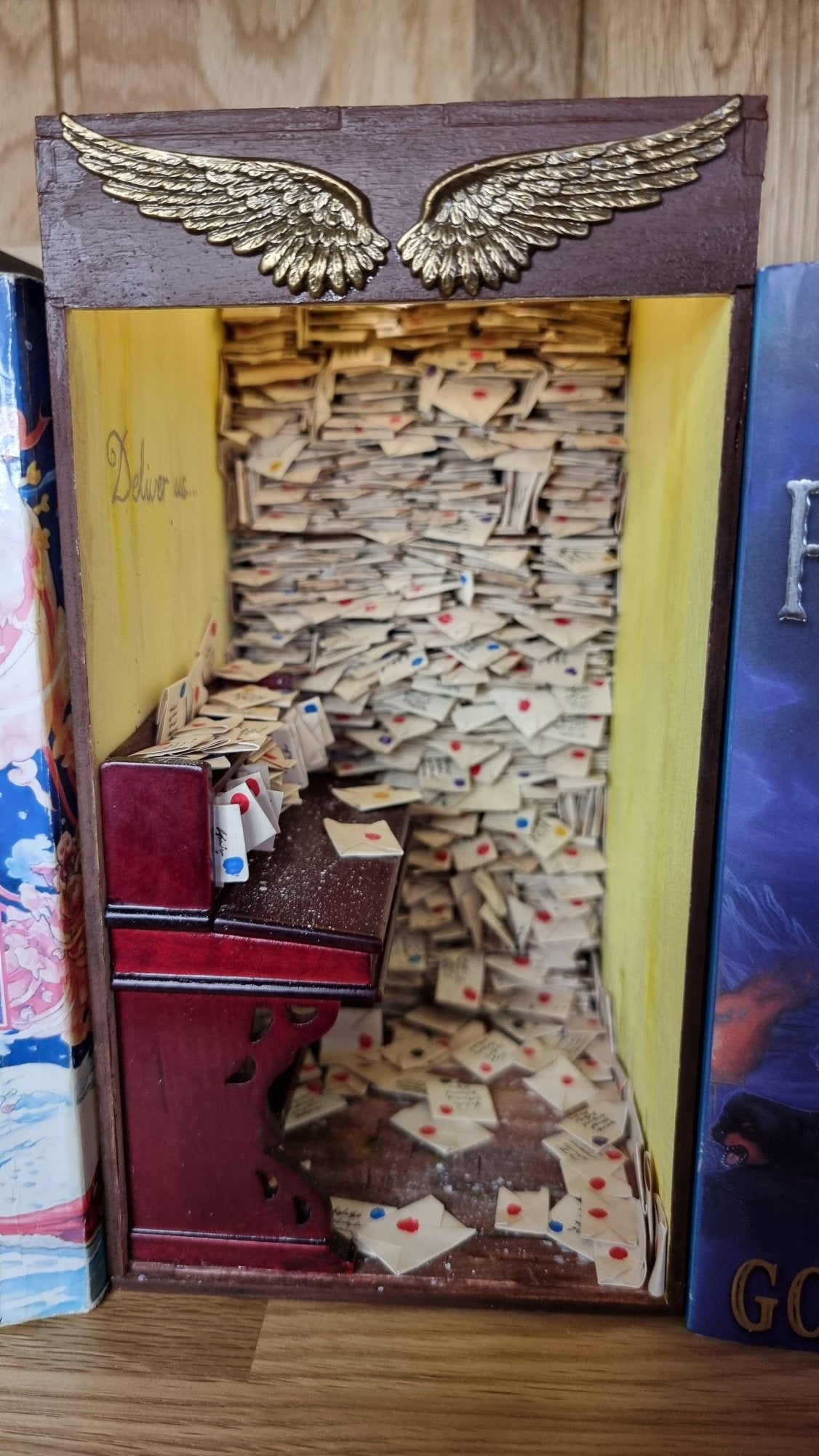 A miniature book nook diorama sits on a shelf beside a copy of Terry Pratchett's Going Postal. The book nook contains a desk littered with letters. Shelves crammed with letters reach from the floor to the roof. The top of the book nook is framed with 2 golden stamped wings. Writing on the yellow wall above the desk reads "Deliver Us"