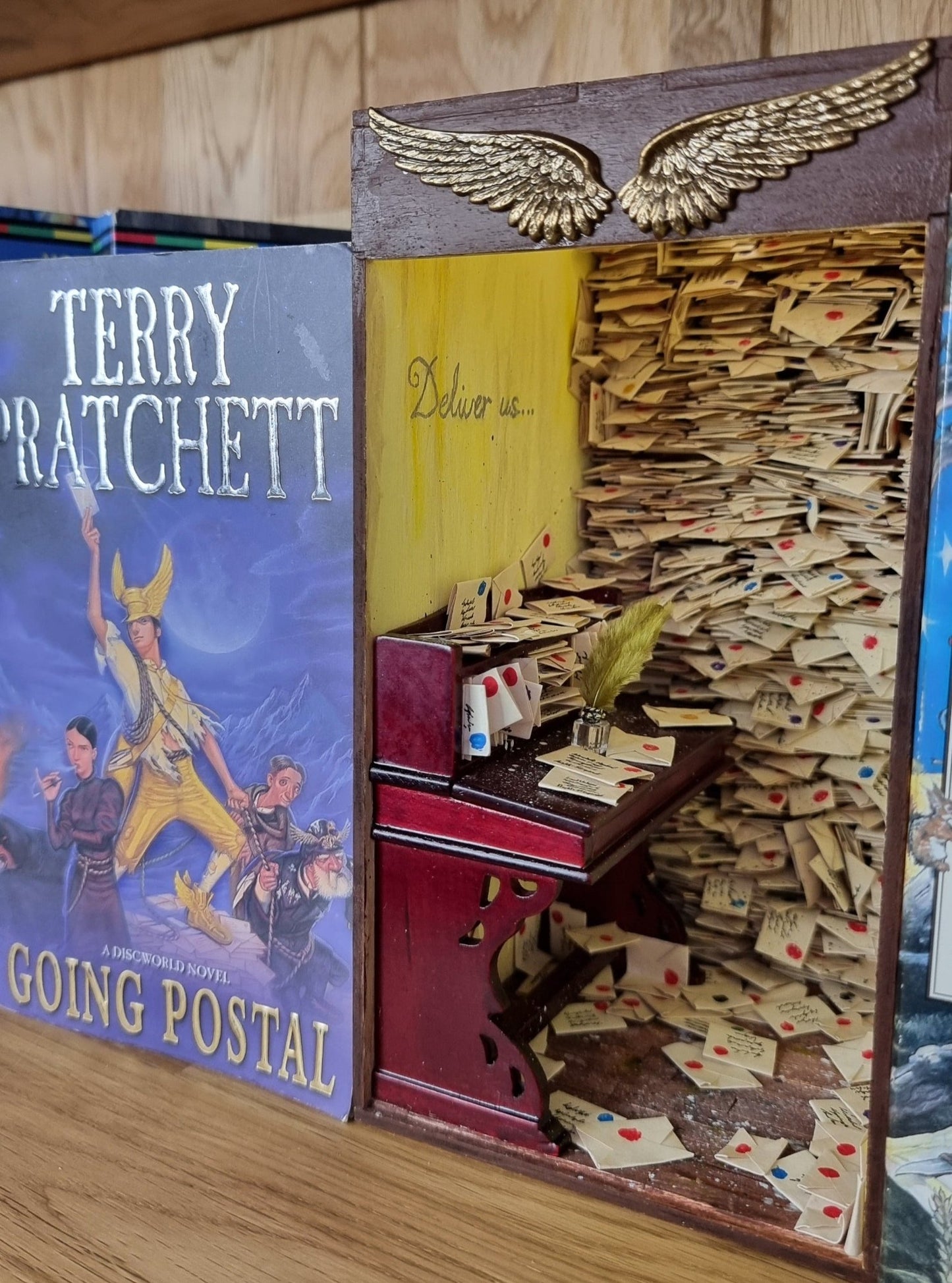 A miniature book nook diorama sits on a shelf beside a copy of Terry Pratchett's Going Postal. The book nook contains a desk littered with letters. Shelves crammed with letters reach from the floor to the roof. The top of the book nook is framed with 2 golden stamped wings