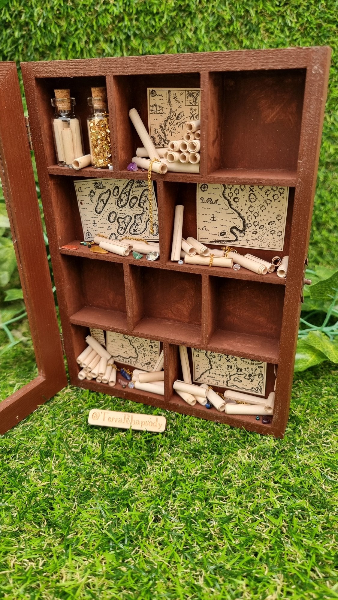 A miniature 1:12 scale tall brown cabinet filled with scrolls of paper sits against a green background. The cabinet has a clear front and 10 shelves or alcoves filled with treasures. 4 alcoves are empty, painted brown. The shelves contain vintage pirate maps, glass vials, chains and gemstones. @Terra Rhapsody is written on wood in front of it