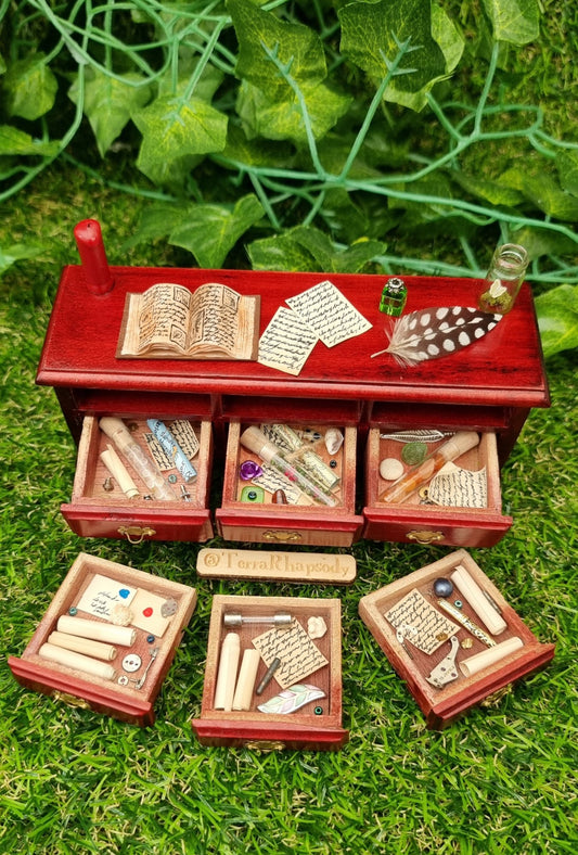 Miniature 1:12 scale dollhouse side board seen from above with 3 open drawers containing bottles parchments and curios. 3 drawers are laid out in front containing parchments,  letters and trinkets. The countertop has a red candle, open book, butterfly bottle, inkwell, quill and parchments 