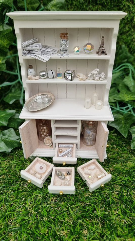 Miniature 1:12 scale white dresser sits against a green background. The dresser has open cupboard doors revealing a white rose and bottles of gemstones. 4 drawers are arranged in front containing light-hued curios and trinkets. The counter top and 2 shelves above contain more candles, crystals, glass bottle, shells, coral and sheet music