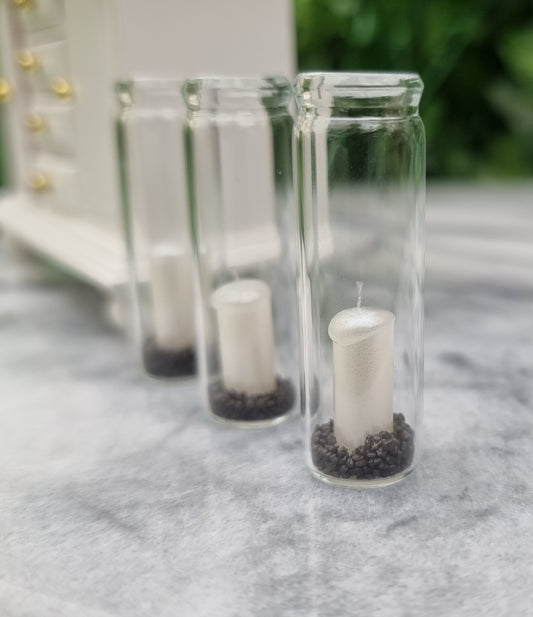 A row of 3 tall 1:12 scale glass hurricane lanterns containing a pearlescent white candles and black sand sit beside a miniature white dresser.