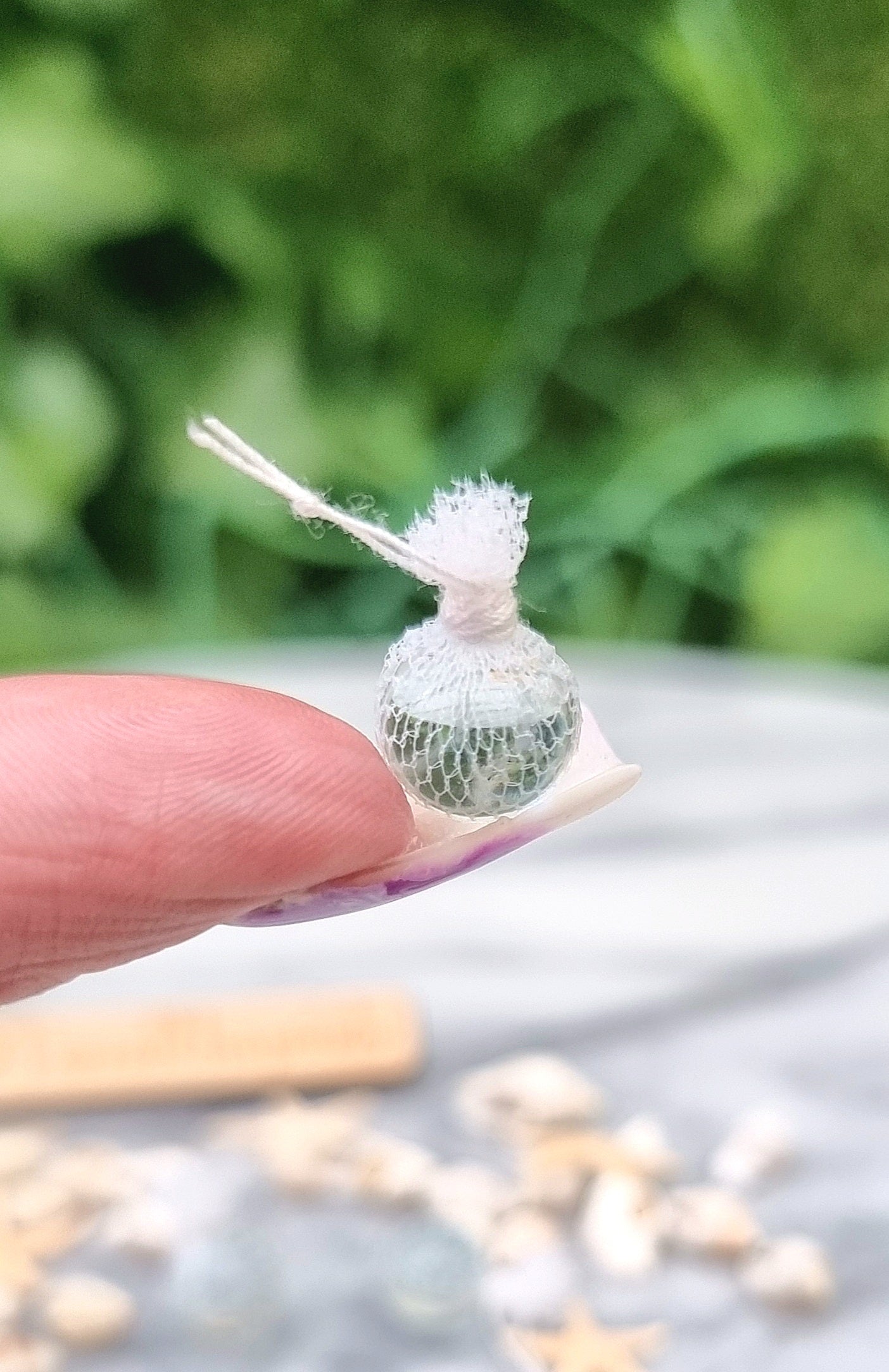 Miniature 1:12 scale clear glass fishing float covered in white netting with a loop of white rope at the top sits on the end of a long fingernail. A slab of white and grey marble sits in the background.