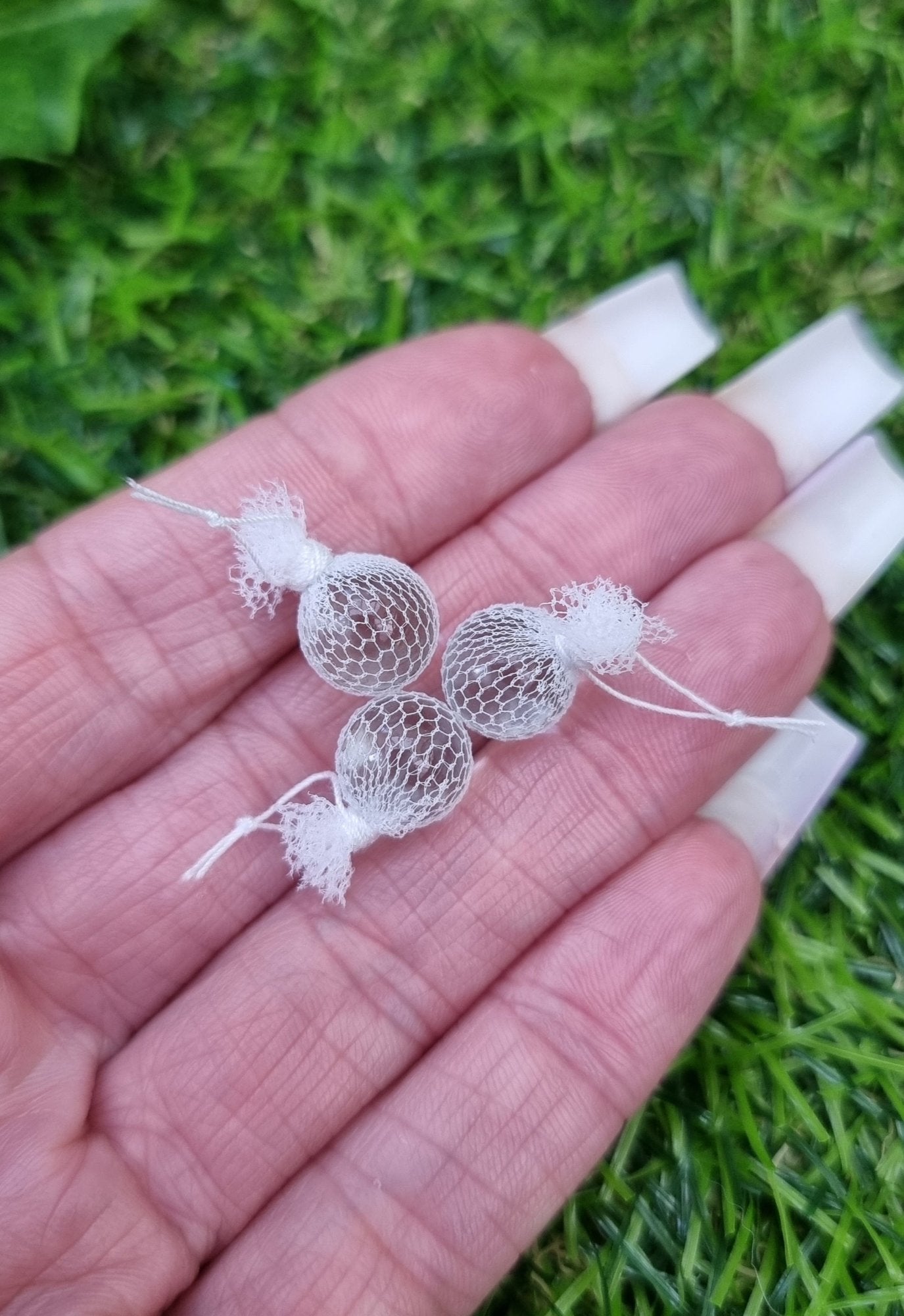 A hand holds 3 miniature 1:12 scale clear glass  fishing floats covered in white netting with a loop of white  rope at the top of each. Held against green background