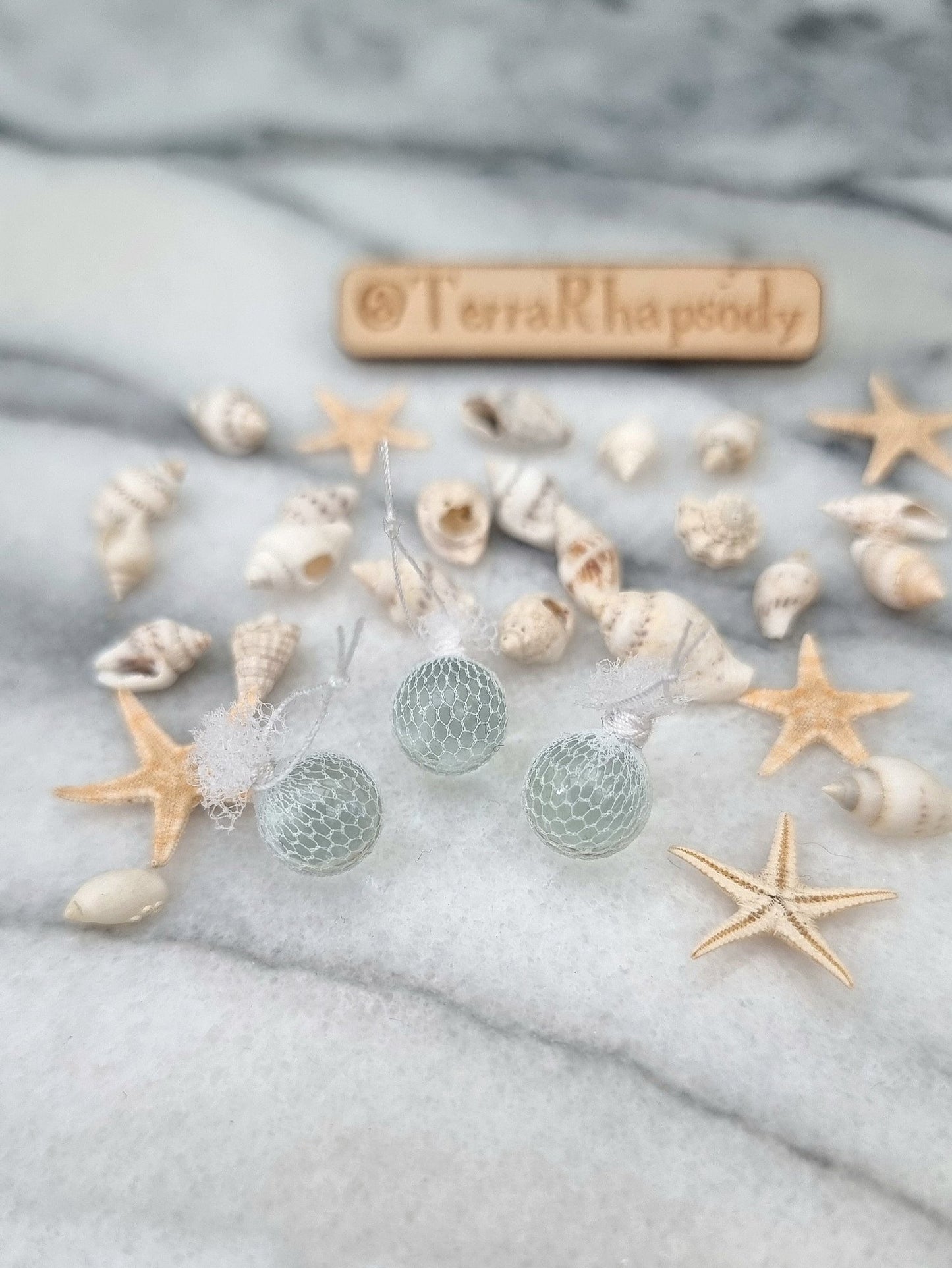 3 miniature 1:12 scale clear glass  fishing floats covered in white netting with a loop of white rope at the top of each. Sat on a white and grey marble background surrounded by shells and starfish. @TerraRhapsody is written on wood in the background