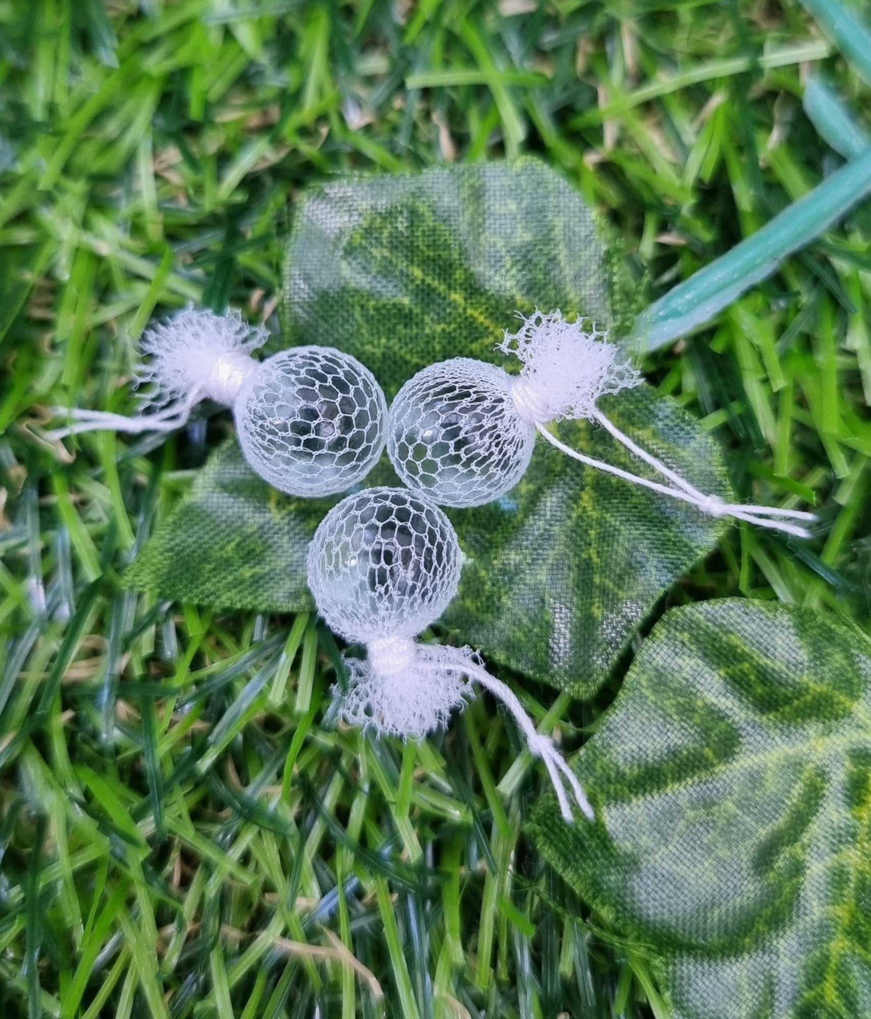 3 miniature 1:12 scale clear glass  fishing floats covered in white netting with a loop of white rope at the top of each. Sat on a green ivy leaf against green backgroun