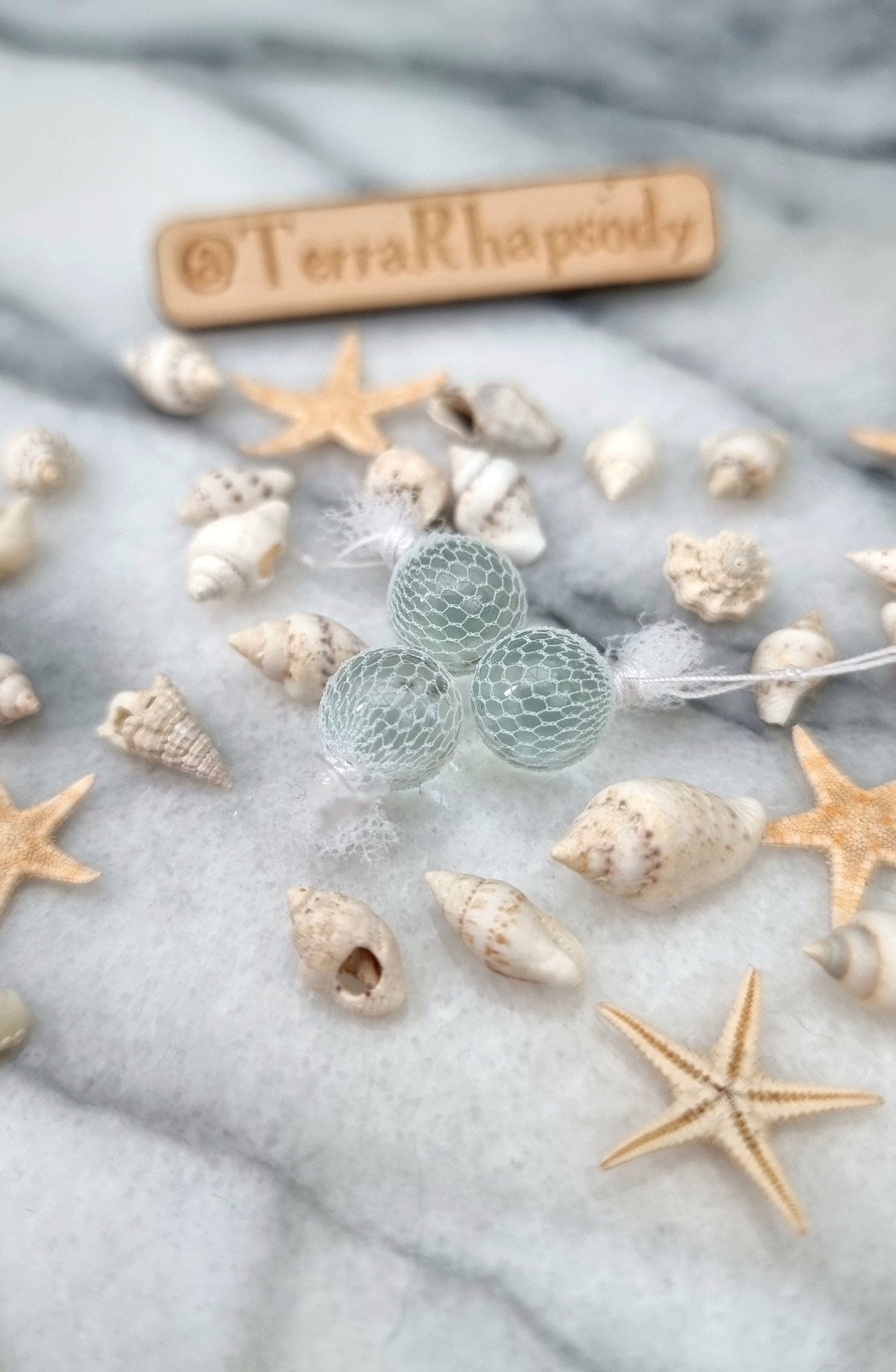 3 miniature 1:12 scale clear glass  fishing floats covered in white netting with a loop of white rope at the top of each. Sat on a white and grey marble background surrounded by shells and starfish. @TerraRhapsody is written on wood in the background