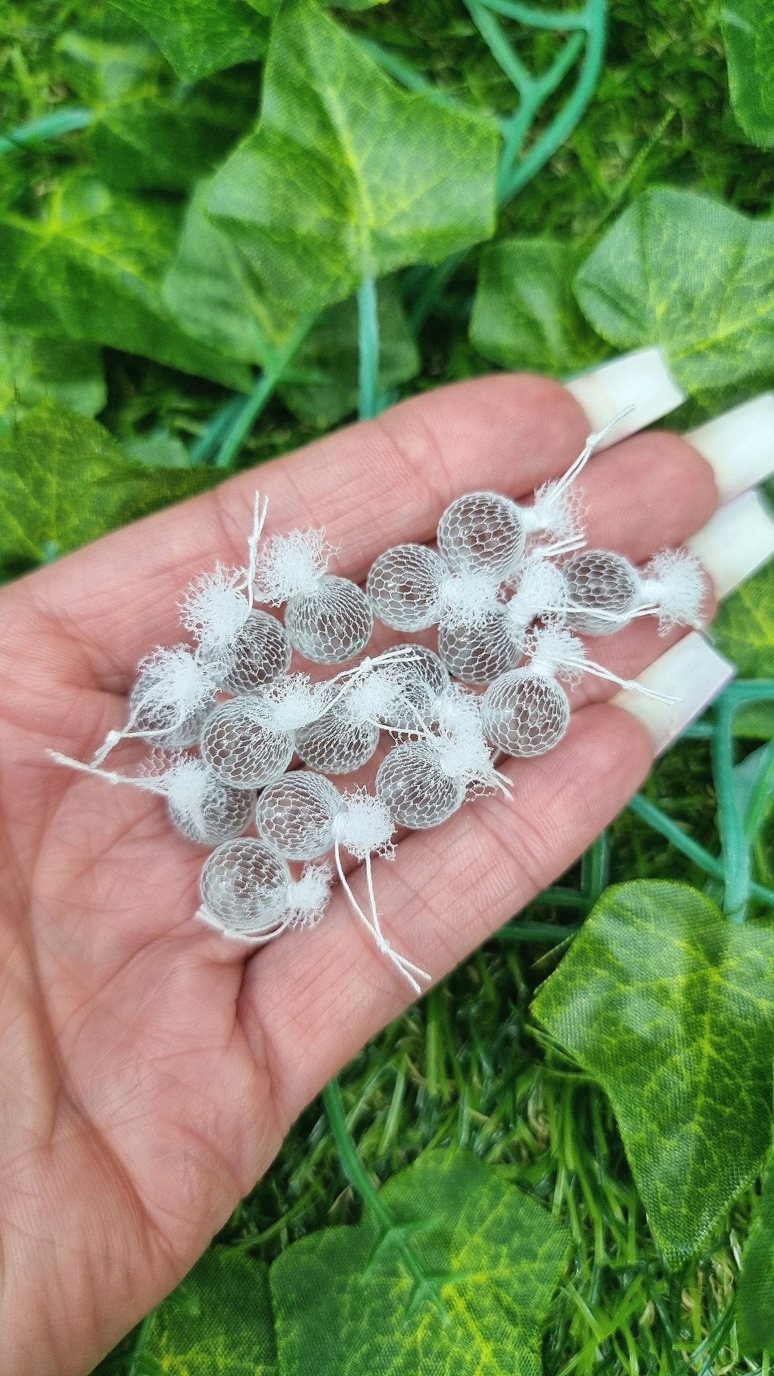 A hand holds 15 miniature 1:12 scale clear glass fishing floats covered in white netting with a loop of white  rope at the top of each. Held against green background