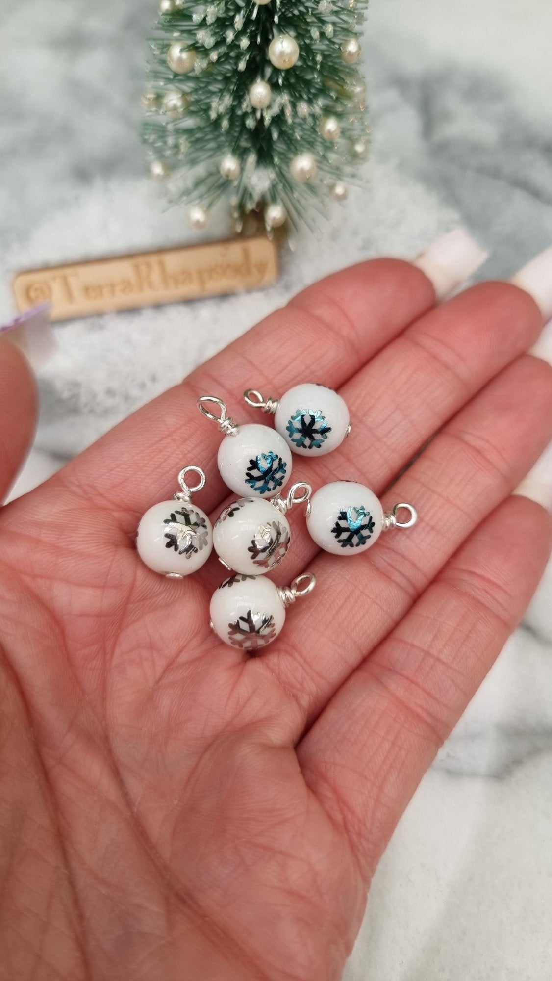 6 Miniature 1:12 Christmas baubles are displayed in an open hand. The baubles are white with silver and blue snowflake motifs. @TerraRhapsody is written on wood in the background