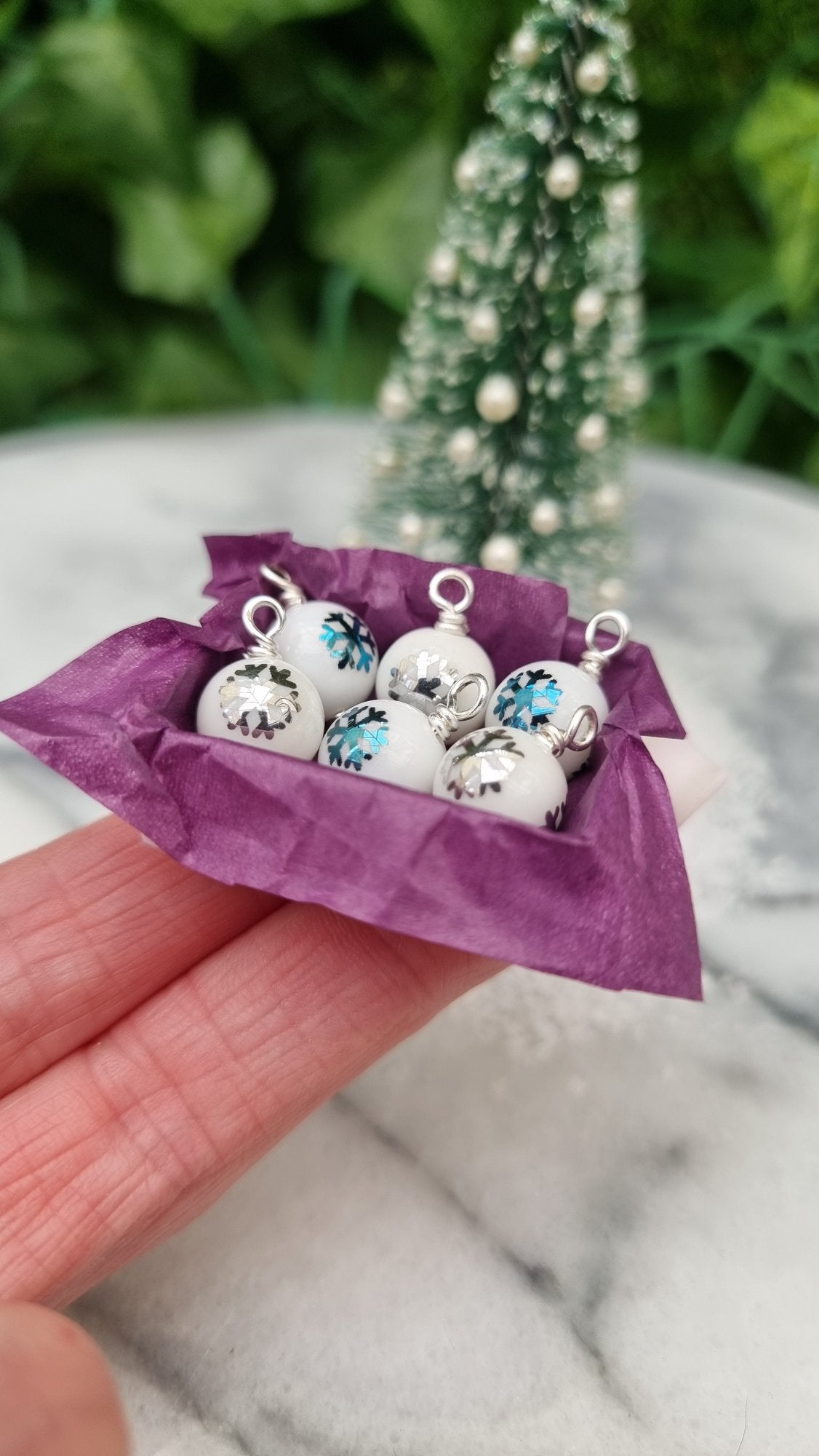 Miniature 1:12 scale open tray box contains 6 Christmas baubles. The baubles are white with silver and blue snowflake motifs. The box is lined with purple paper, displayed on two fingers in front of a Christmas tree