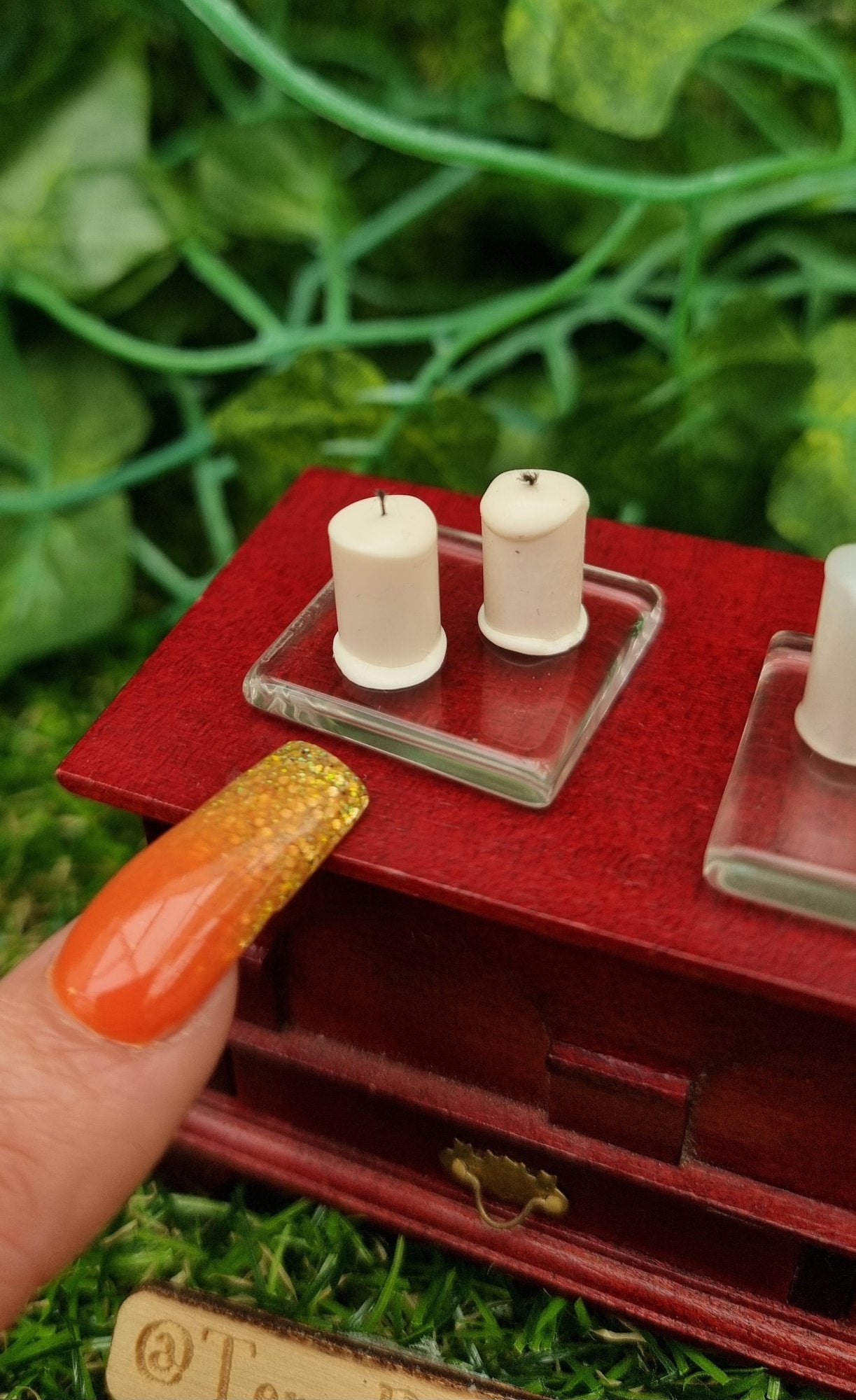 A finger points at 1:12 scale miniature ivory candles sitting on a square glass plate on top of a wooden chest. There are 2 ivory candles on the glass plate . @TerraRhapsody is written on wood in the foreground